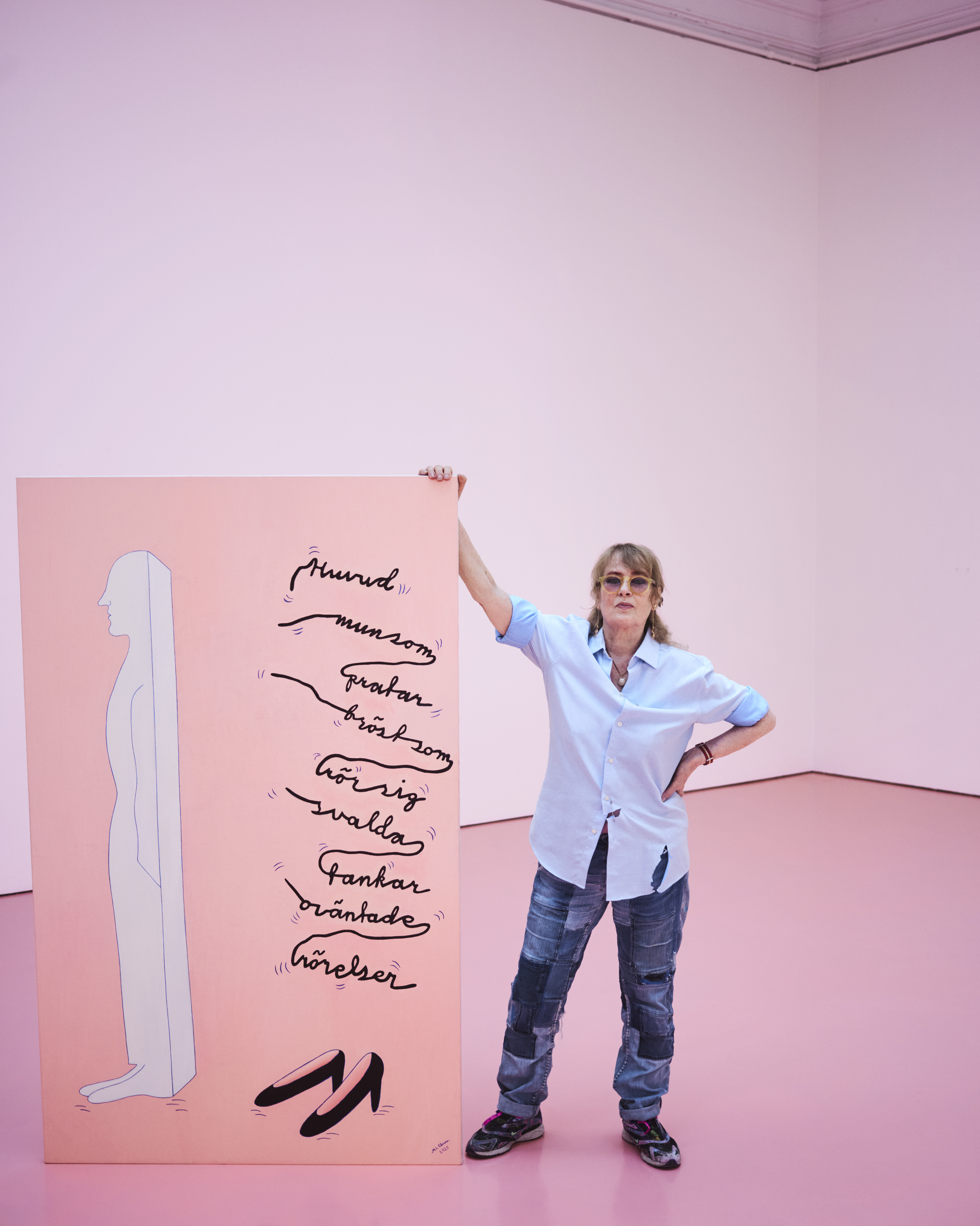 En person i vardagskläder står i ett rosa rum bredvid ett stort konstverk med en siluett, text och tecknade skor.