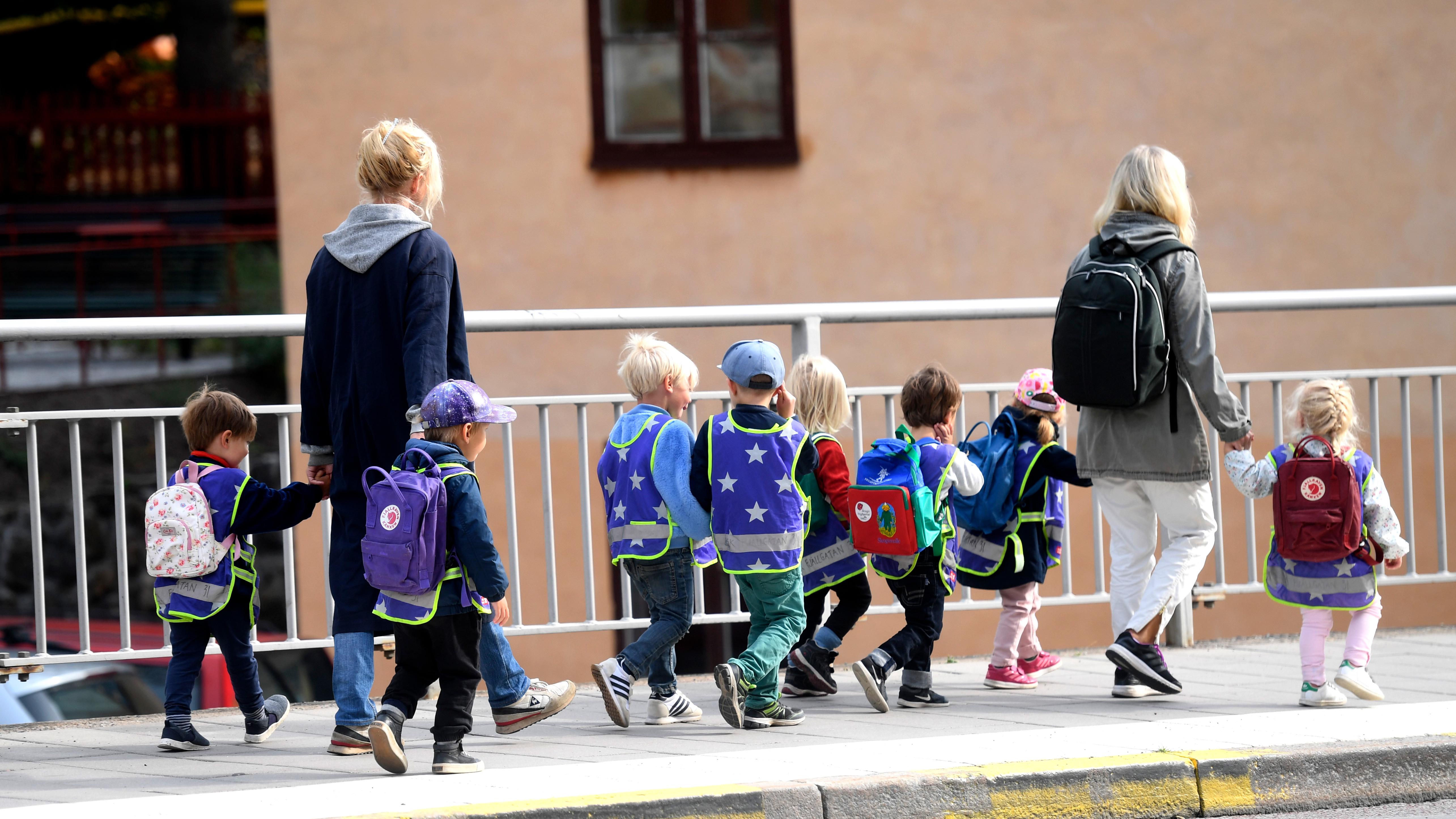 Två vuxna övervakar en grupp barn med ryggsäckar och reflexvästar som går på en trottoar bredvid ett metallräcke.