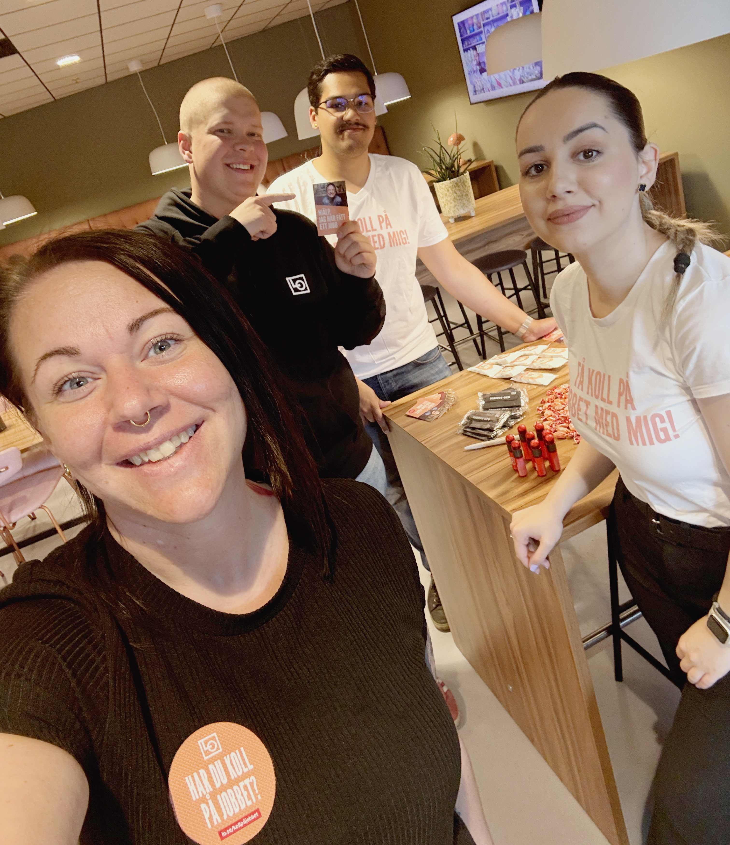 Fyra personer står vid ett bord med reklammaterial, två har likadana tröjor och en håller i ett kort, alla ser mot kameran.