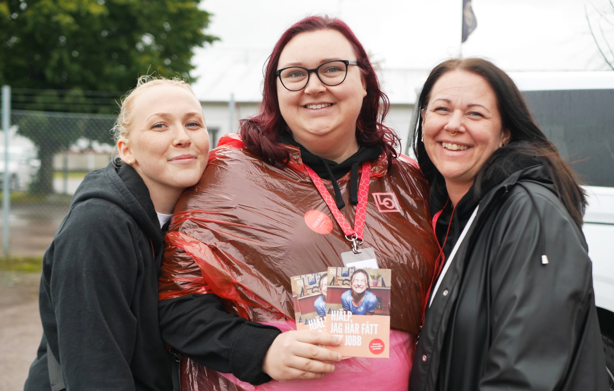 Tre kvinnor står utomhus och tittar mot kameran. En har en röd poncho och håller i ett häfte. Staket och träd syns bakom.