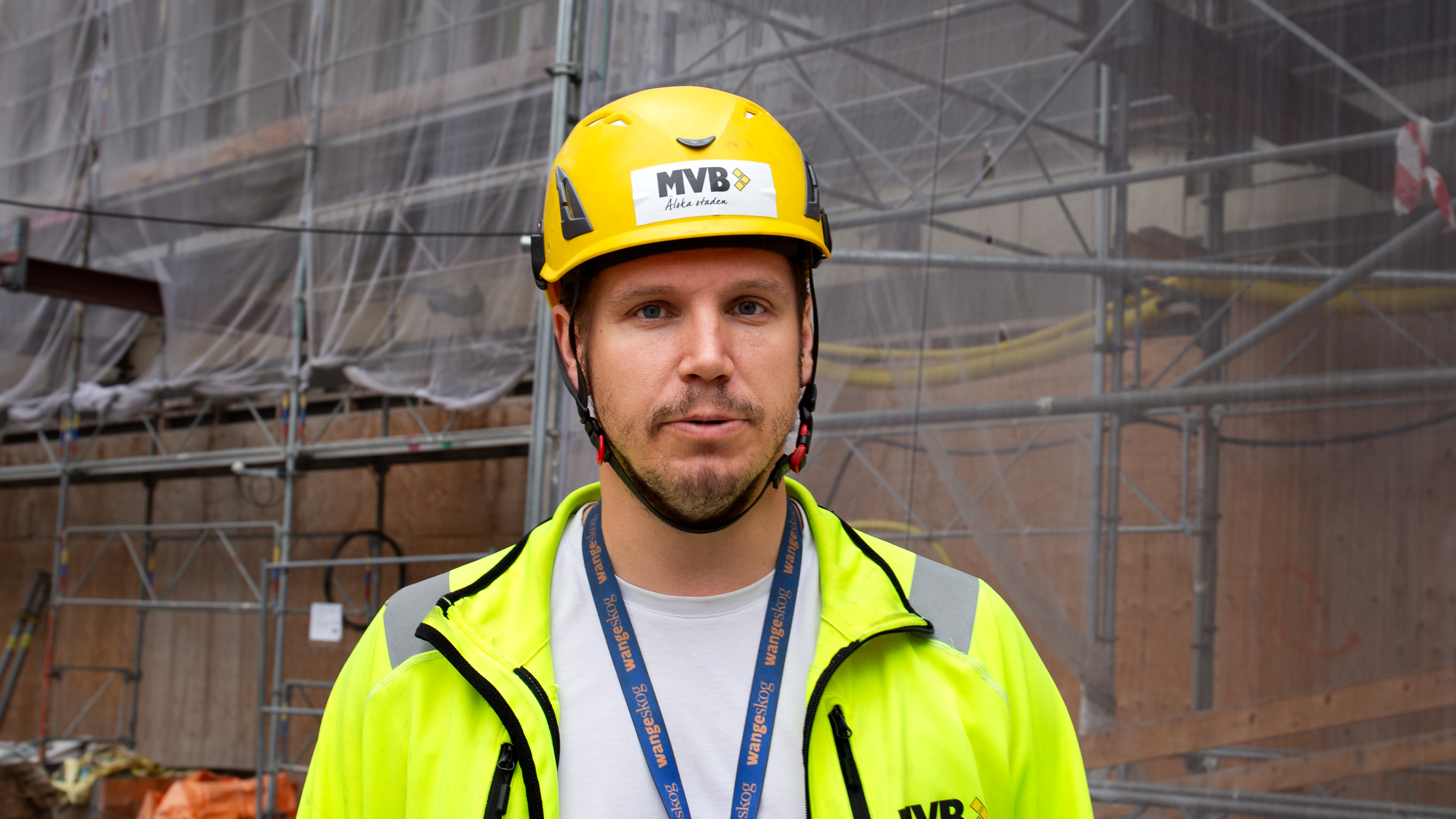 En byggarbetare med gul hjälm och reflexjacka står framför byggnadsställningar på en byggarbetsplats.