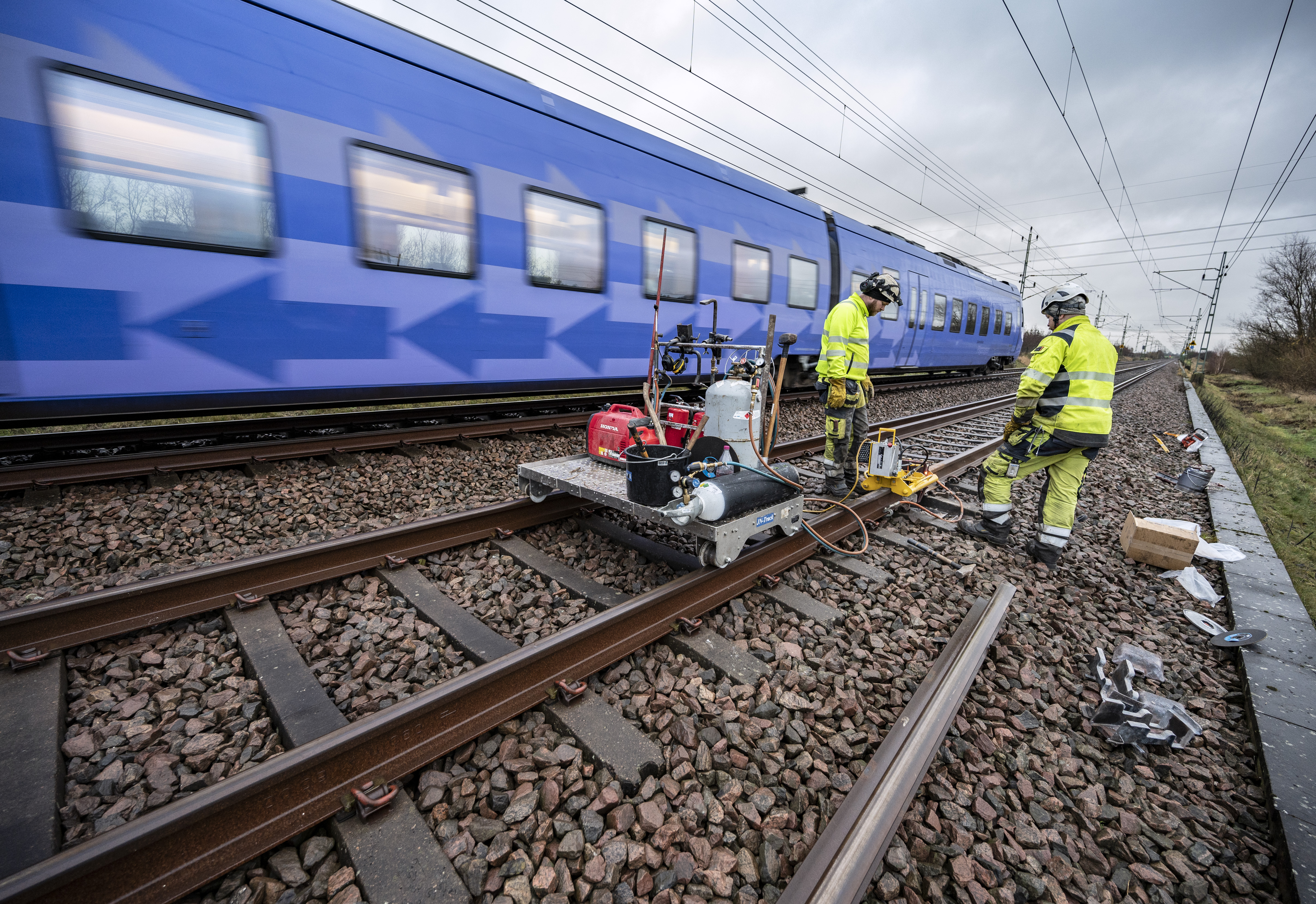 Två arbetare i varselkläder utför underhåll på järnvägsspår medan ett passagerartåg kör förbi på ett intilliggande spår.