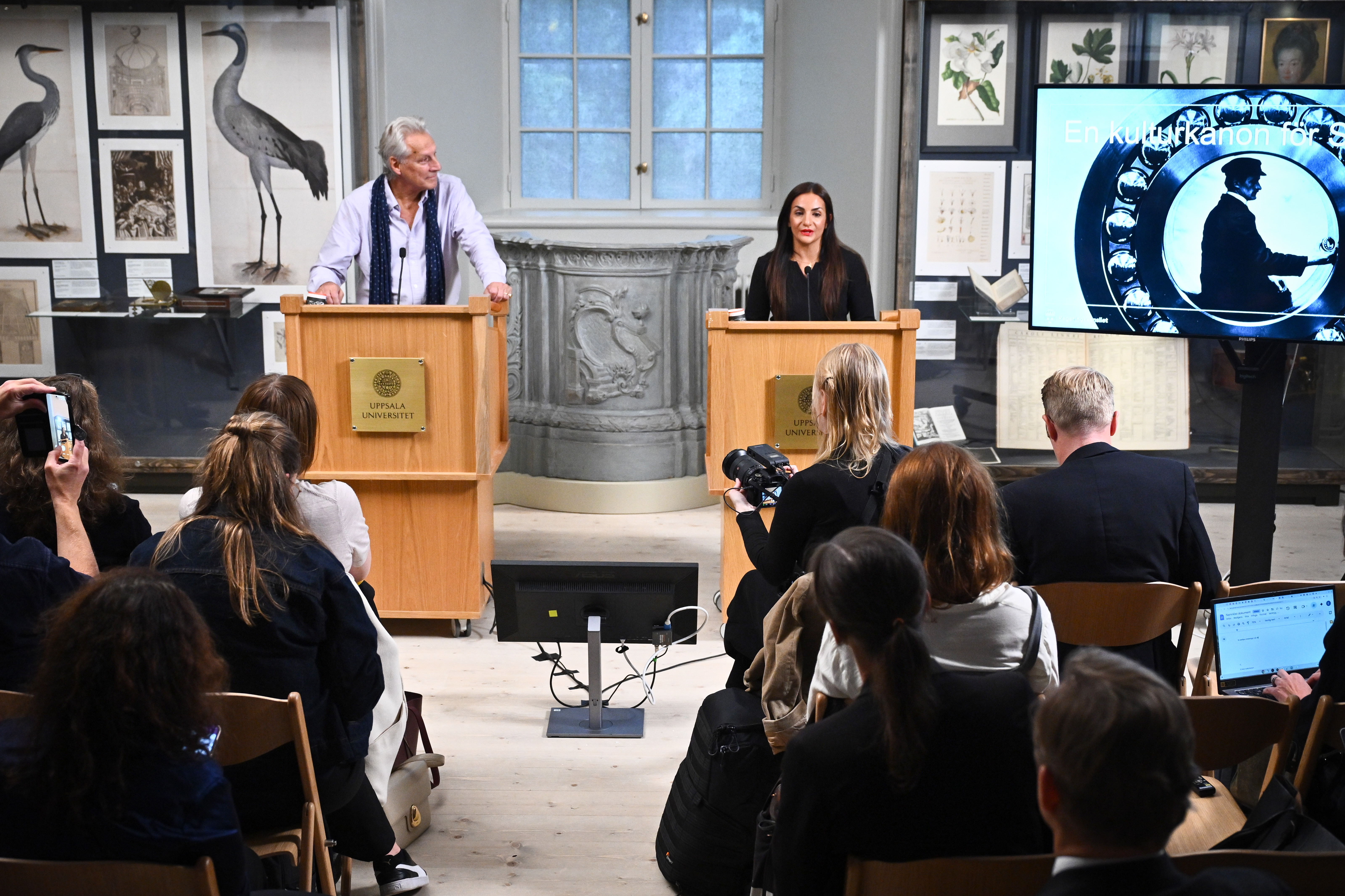 En man och en kvinna står vid talarstolar och talar till en publik i ett rum med fågelbilder och en projicerad bild på skärm.