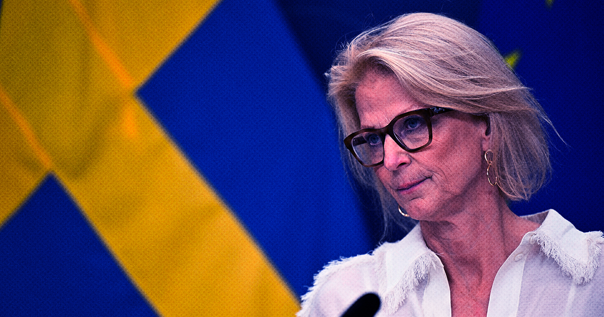 Elisabeth Svantesson, med glasögon och vit skjorta står framför svenska och Europeiska unionens flaggor.