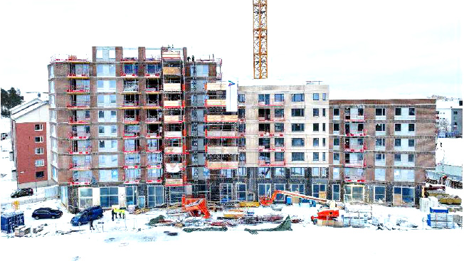 Ett flerfamiljshus under byggnation på vintern, med ställningar, byggutrustning och arbetare på snötäckt mark.