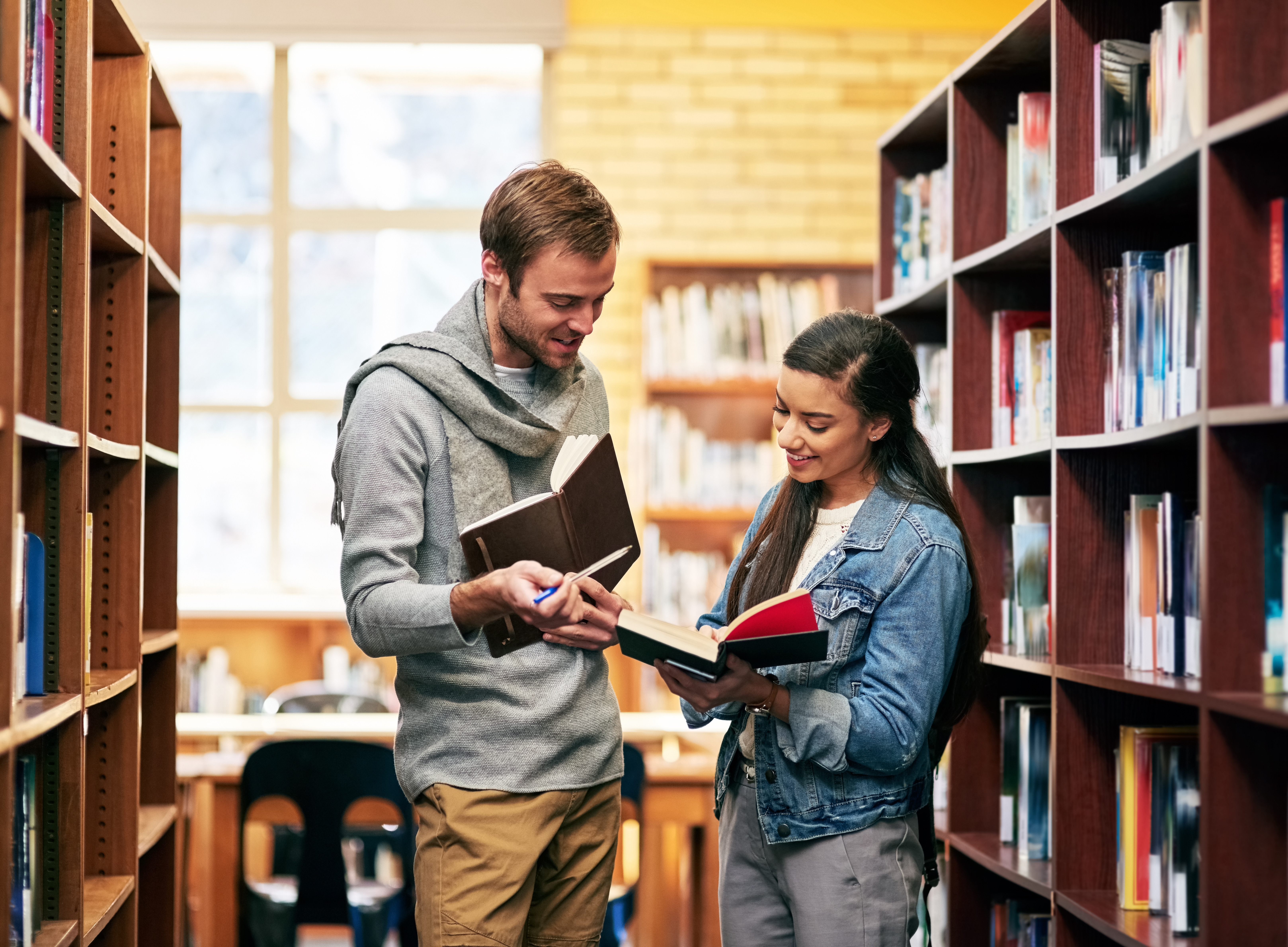 Två personer står mellan bokhyllor i ett bibliotek, håller öppna böcker och pratar med varandra.