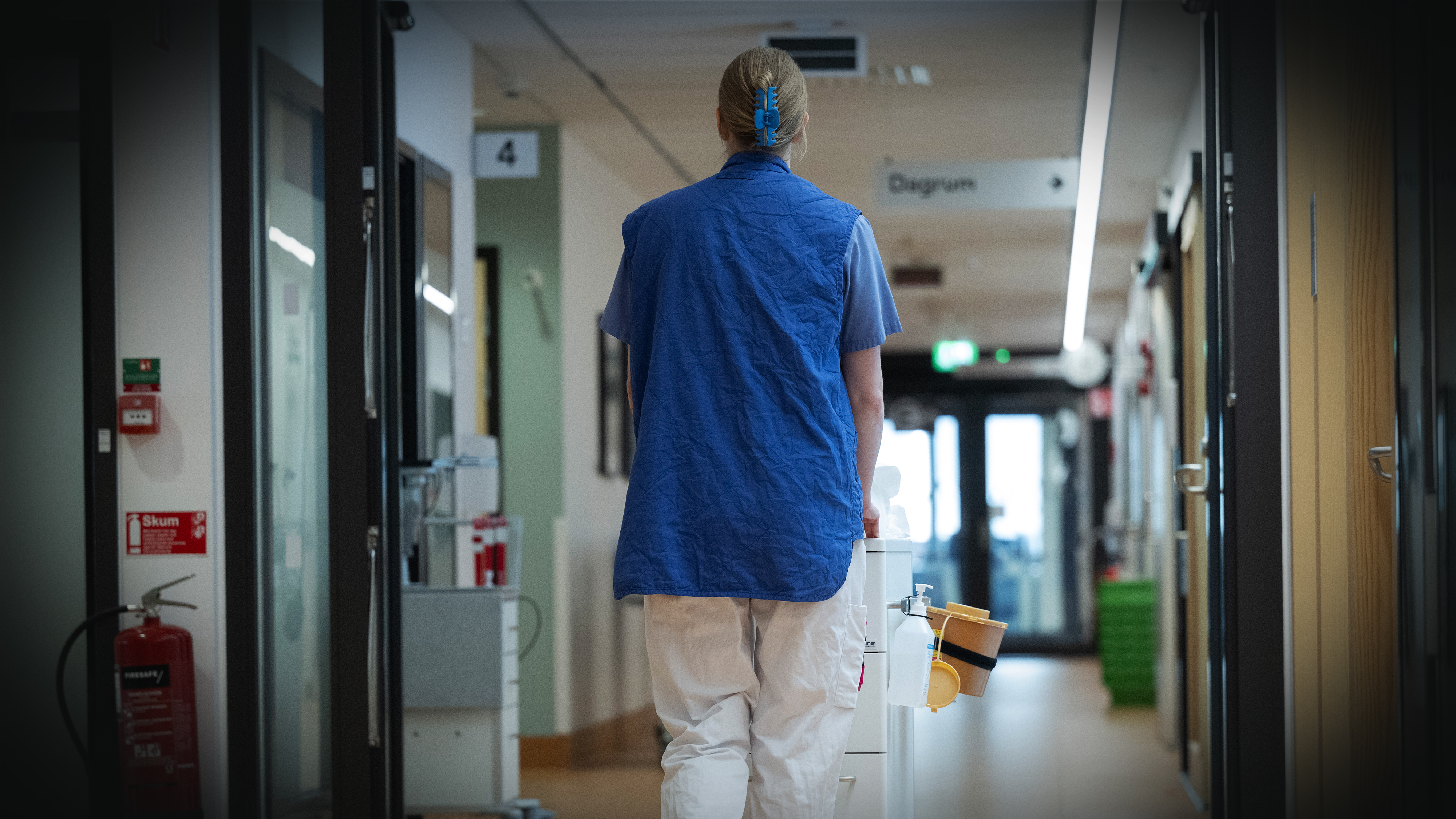En undersköterska går bort från kameran i en sjukhuskorridor med medicinsk utrustning.