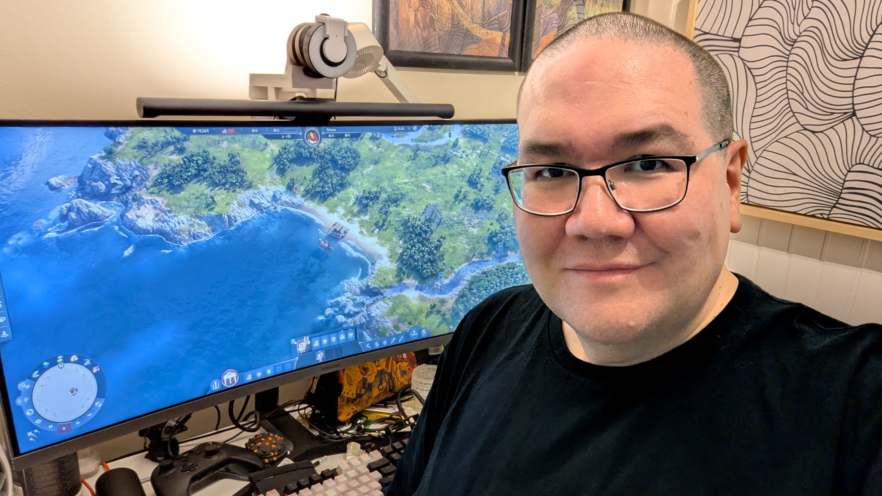 En person med glasögon och svart tröja ler mot kameran framför en datorskärm som visar ett kustlandskap från ett videospel.