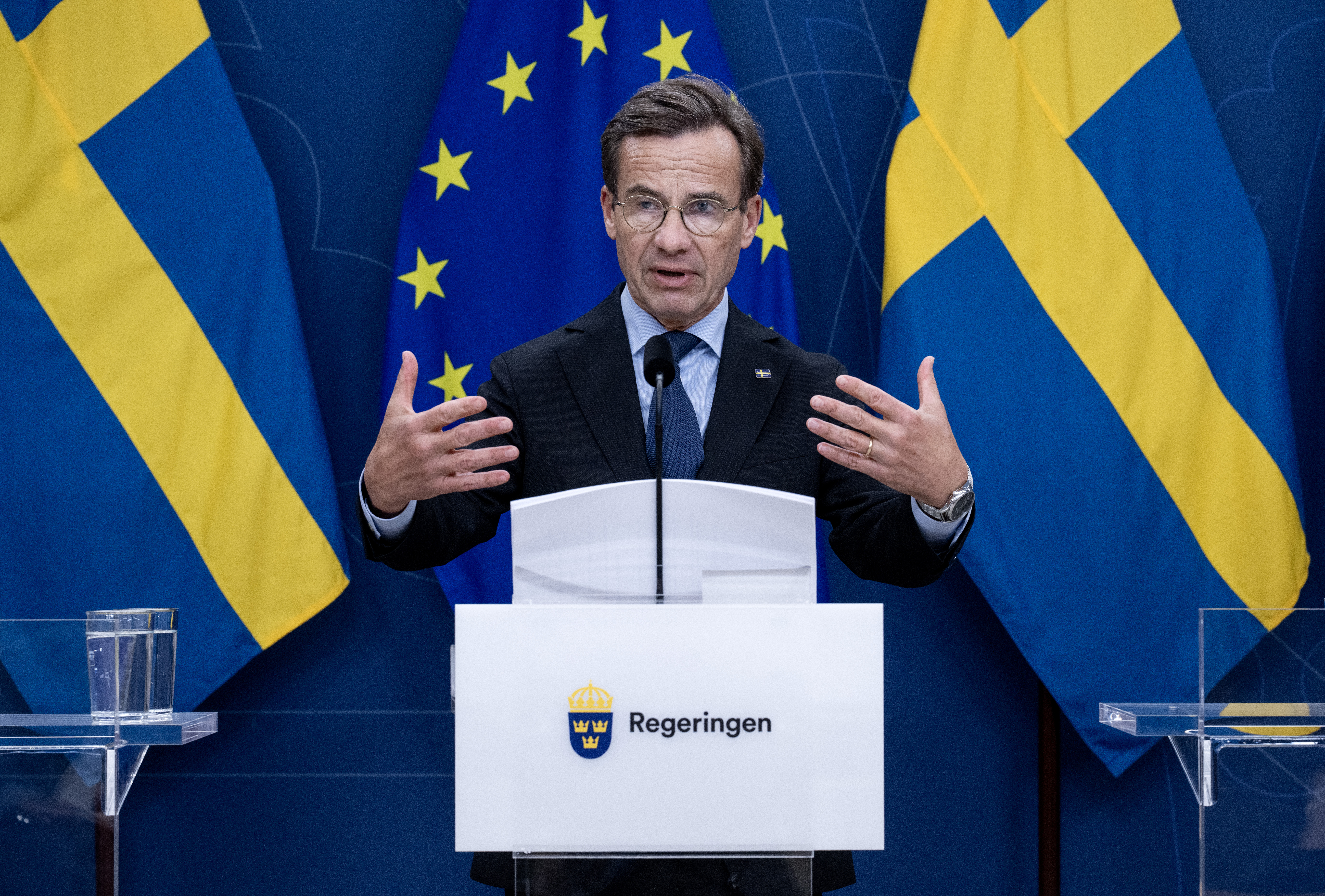 Ulf Kristersson talar vid en talarstol märkt "Regeringen" med svenska och EU:s flaggor i bakgrunden.