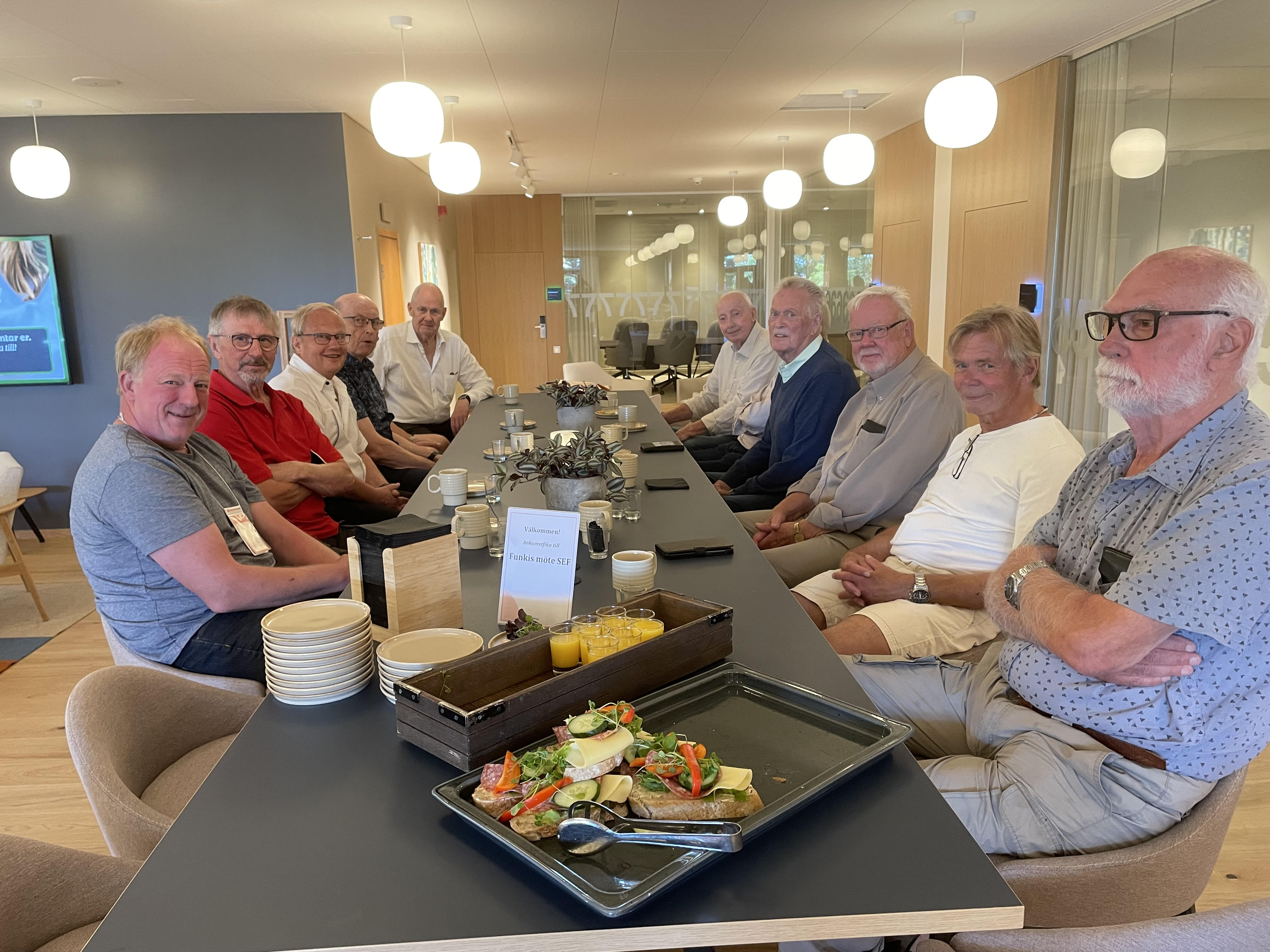 Elva äldre män sitter runt ett långt bord i ett mötesrum med snacks, drycker och tallrikar framför sig.