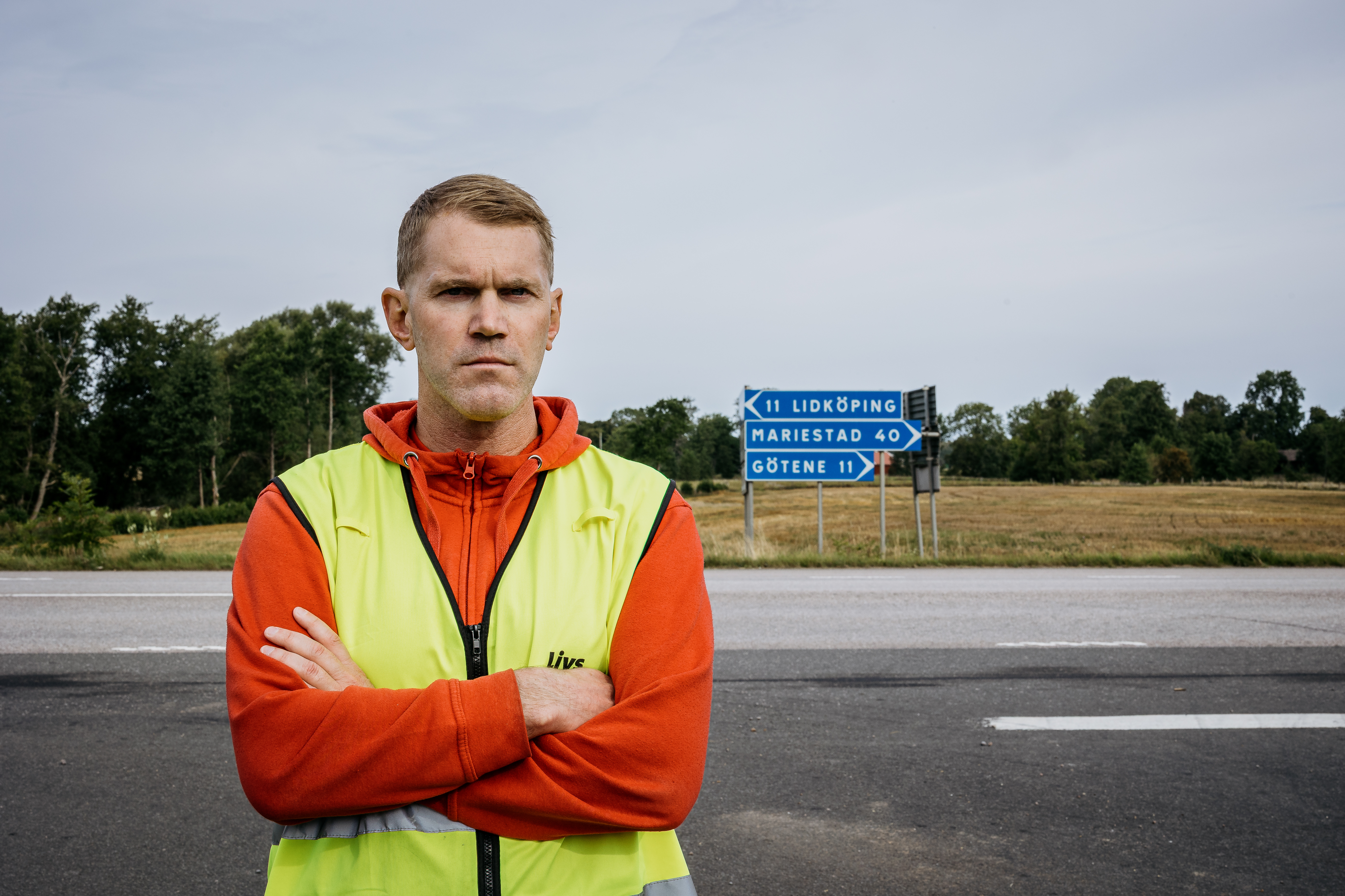 Thomas Nilsson med gul säkerhetsväst står med armarna i kors vid en landsväg, vägskyltar till Lidköping, Mariestad och Götene syns bakom.