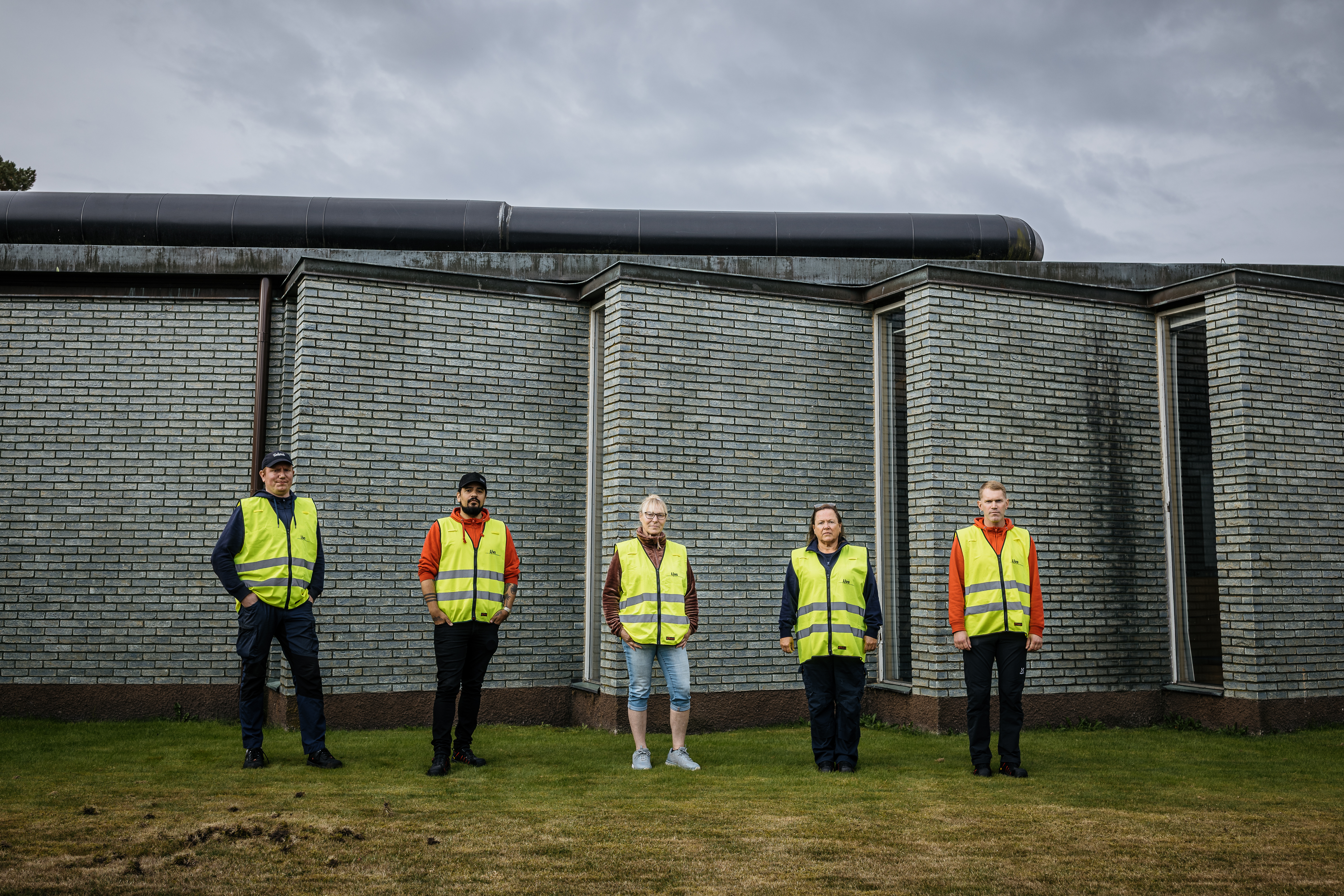Fem personer (Johan Ahlén, David Karlsson, Annica Andersson, Eva Wenne och Thomas Nilsson) med gula varselvästar står på gräs i en rad framför en byggnad av grå tegel under en molnig himmel.