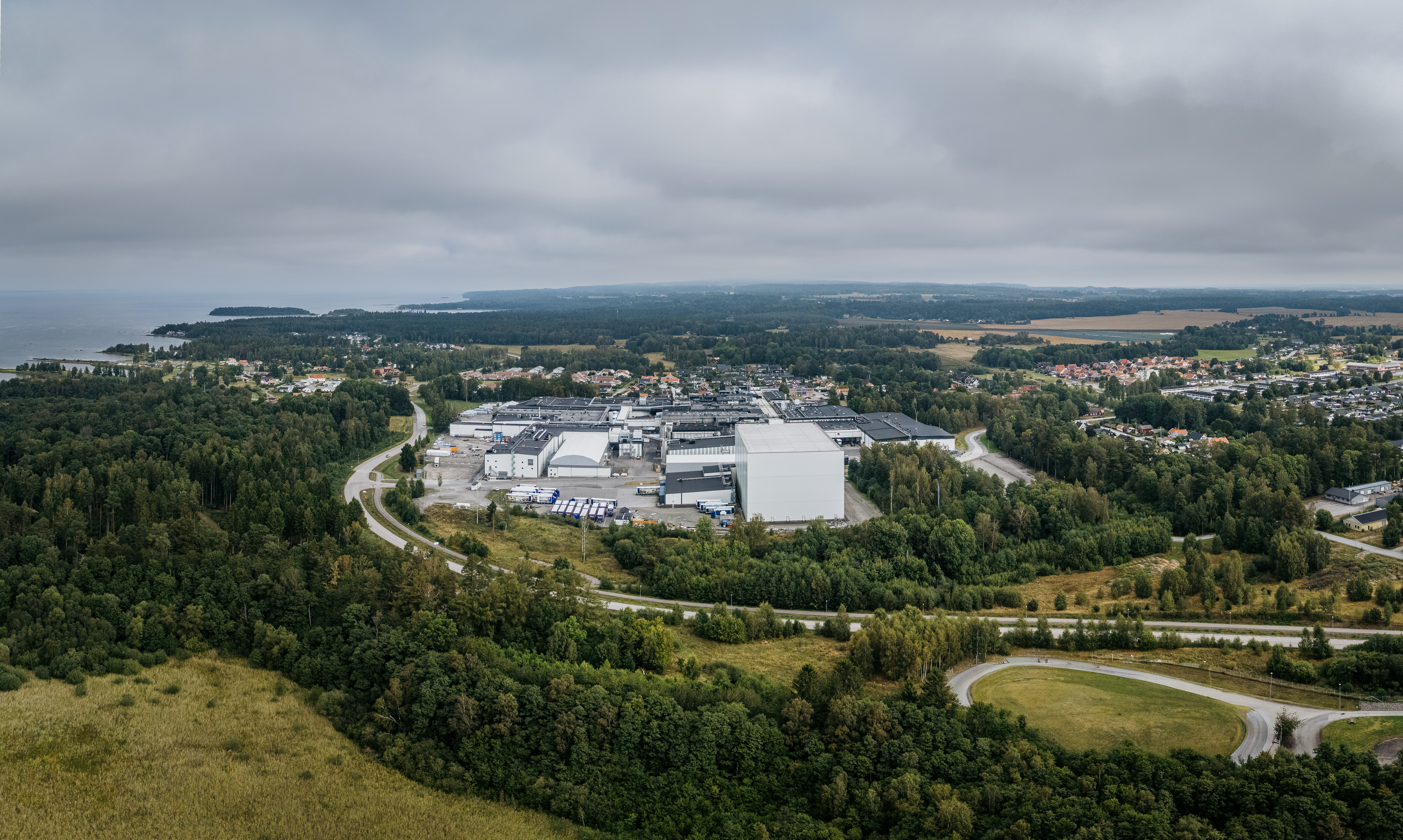 Dafgårds industribyggnad sett uppifrån med omgivande skog, fält och en väg, bostadsområden och kust syns längre bort under molnig himmel.