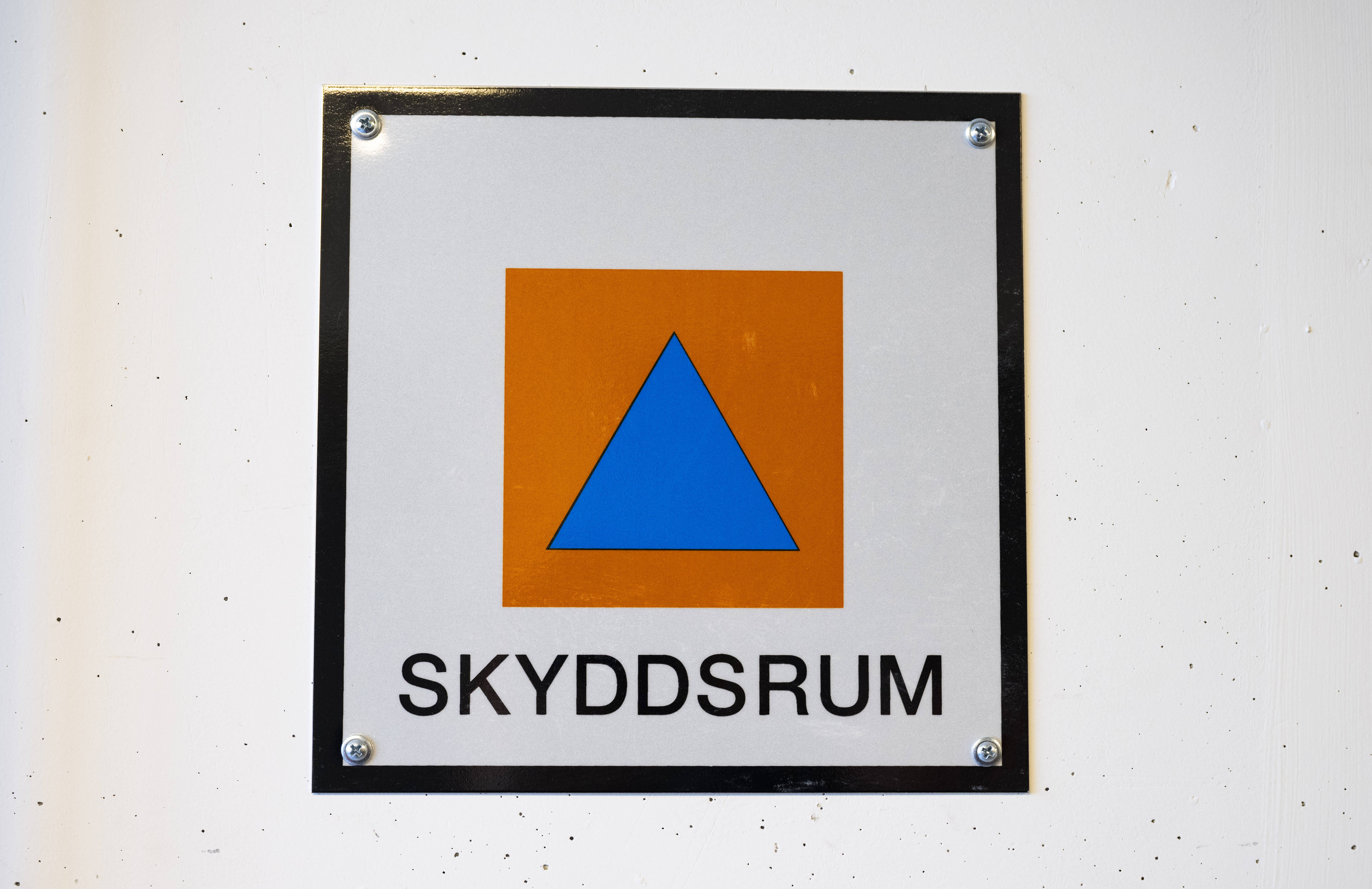 En skylt med ett blått triangel inuti en orange kvadrat och texten "SKYDDSRUM" på vit bakgrund, markerar svenskt skyddsrum.