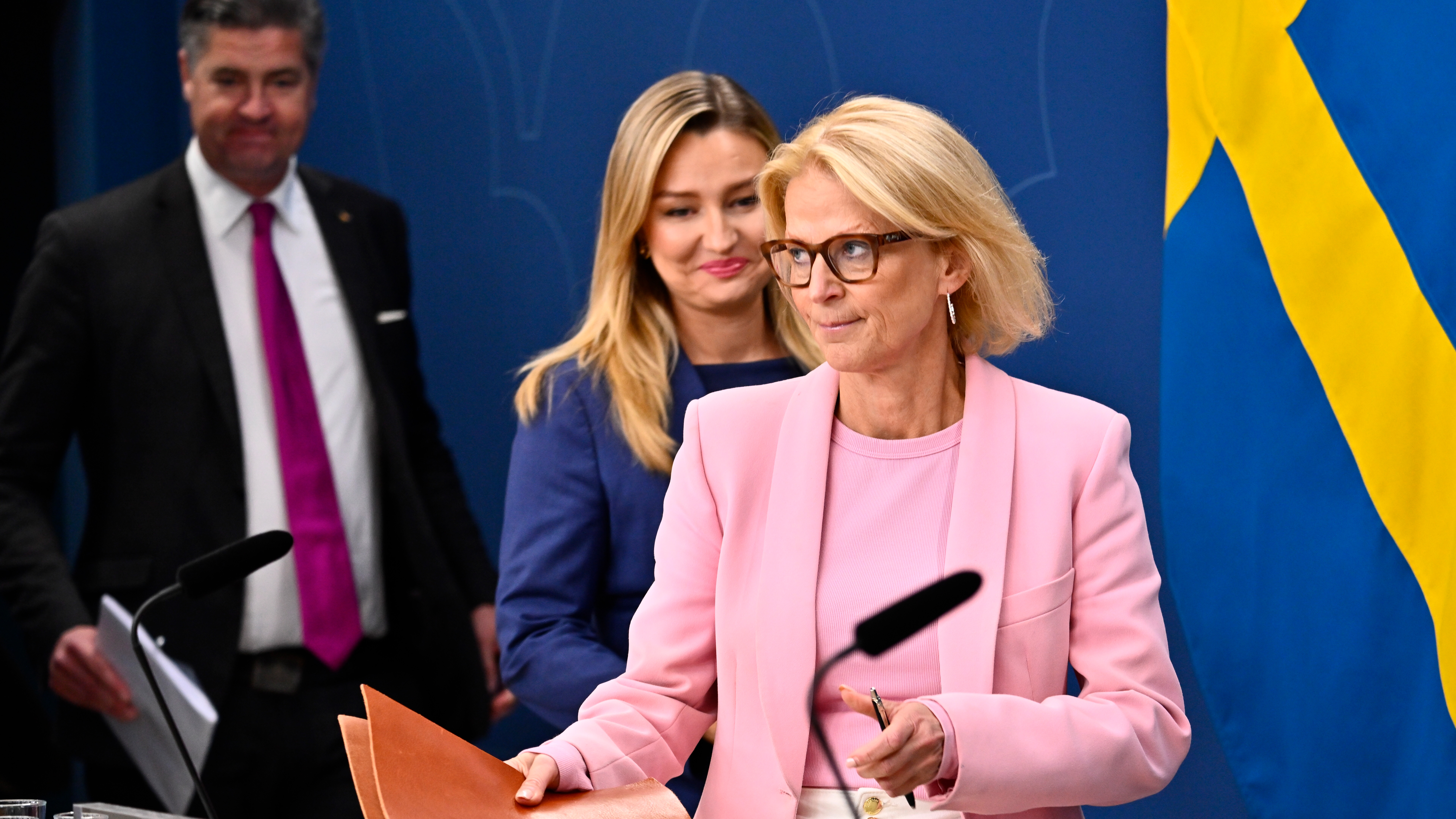 Tre personer står framför en blå vägg och en svensk flagga. Kvinnan längst fram håller papper, har glasögon och ljusrosa kavaj.