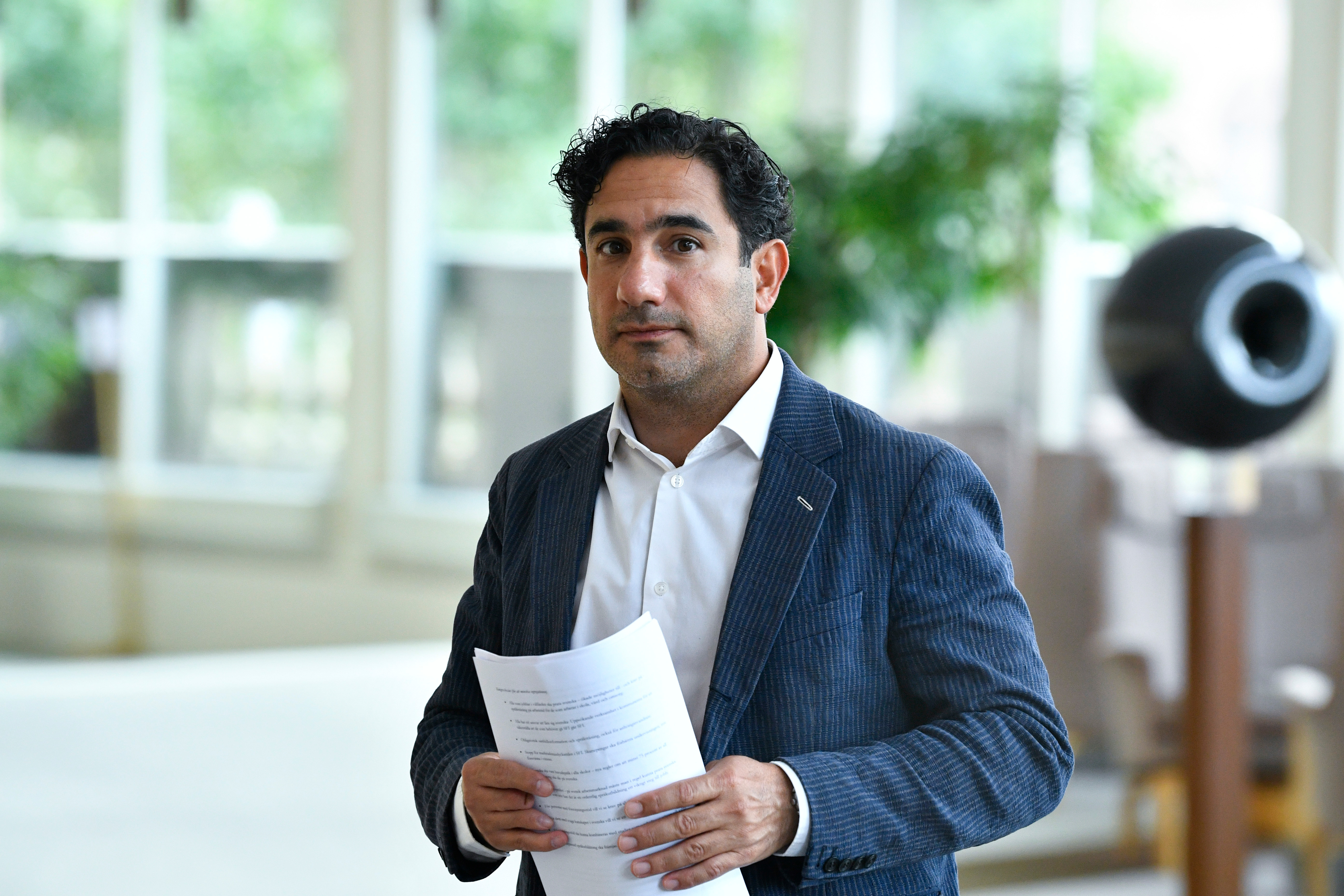 En man i kritstrecksrandig kostym håller papper och tittar mot kameran i en inomhusmiljö med stora fönster och växter.