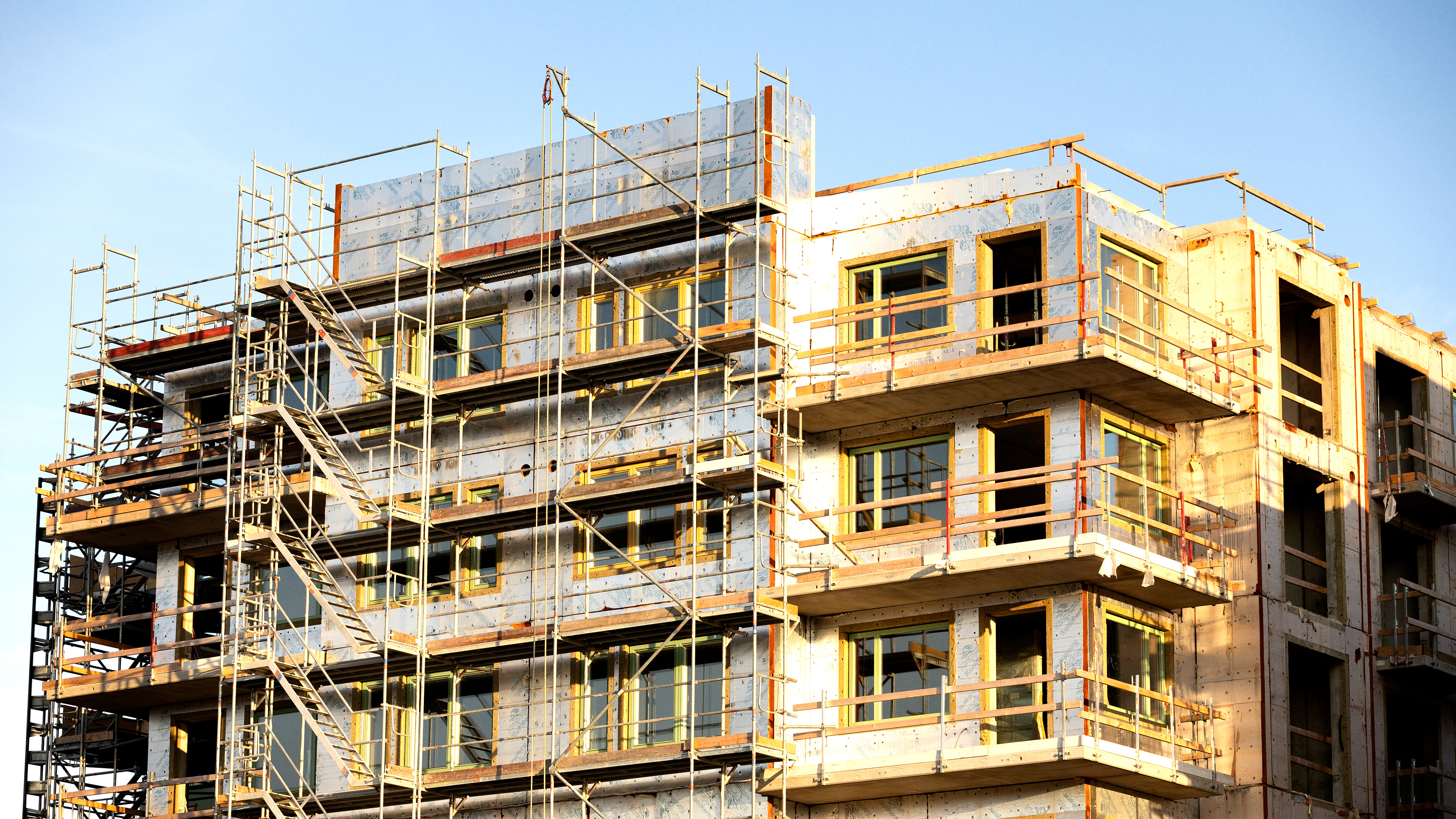 En flervåningsbyggnad under konstruktion är täckt av byggställningar, med synlig isolering och oavslutade ytor.