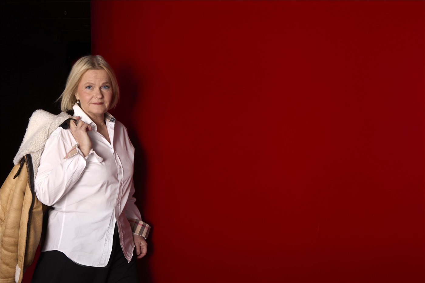 Sissela Kyle står mot en röd vägg och håller en beige kappa över axeln.