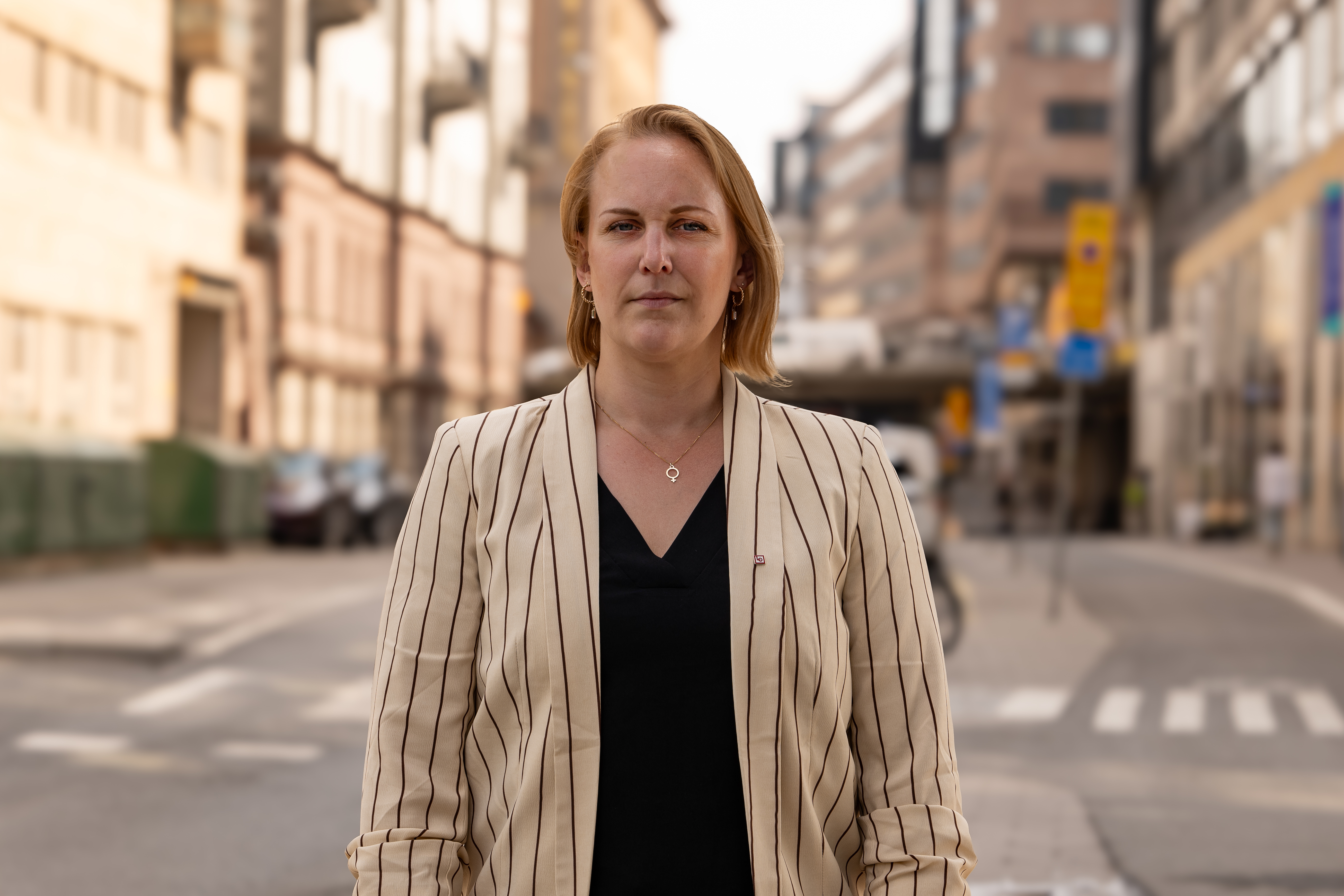 Louise Olsson med randig kavaj står på en stadsgata med byggnader och bilar i bakgrunden.