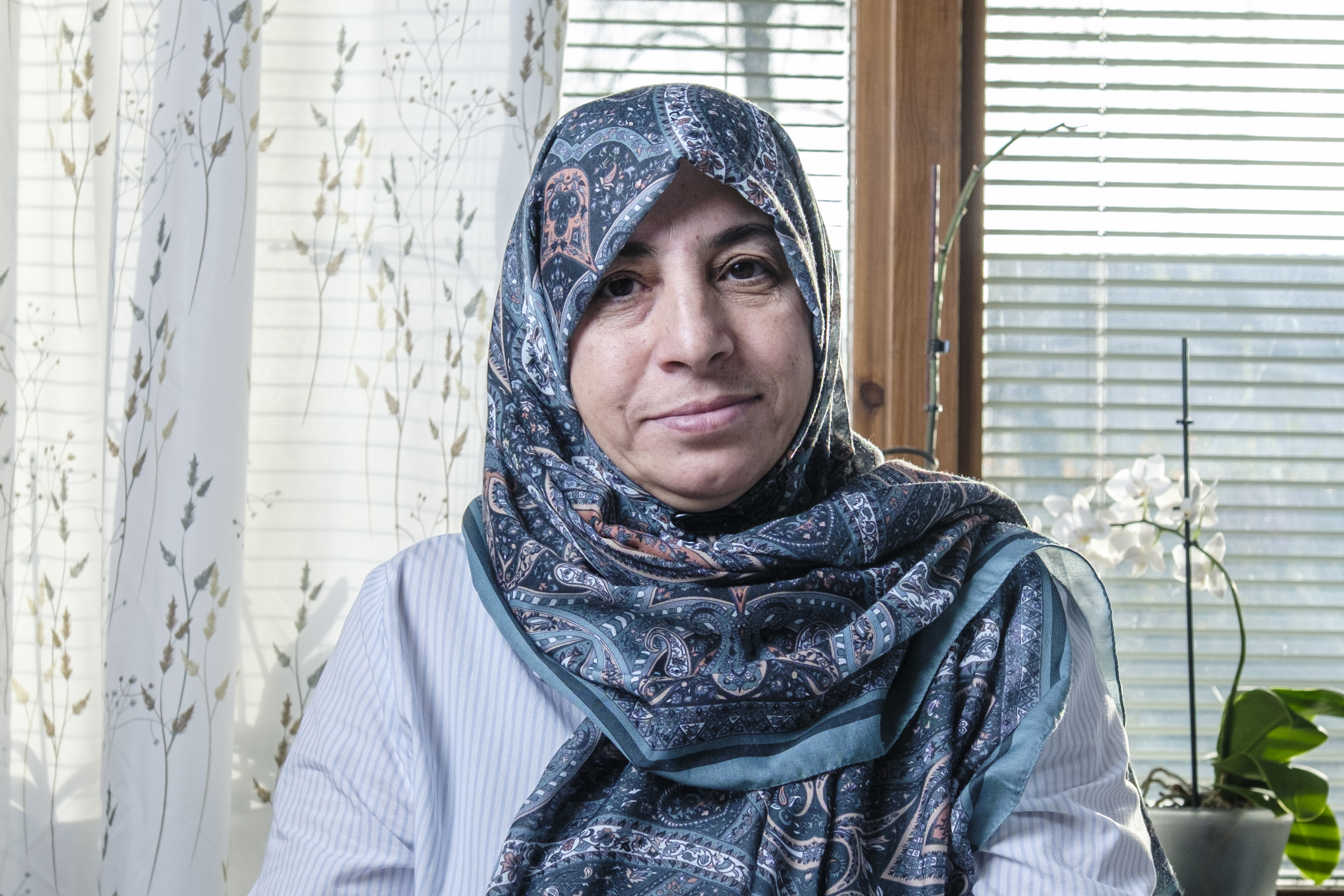Iförd en mönstrad sjal och en ljus skjorta sitter Meryem inomhus vid ett fönster med persienner och gardiner, med växter i bakgrunden.
