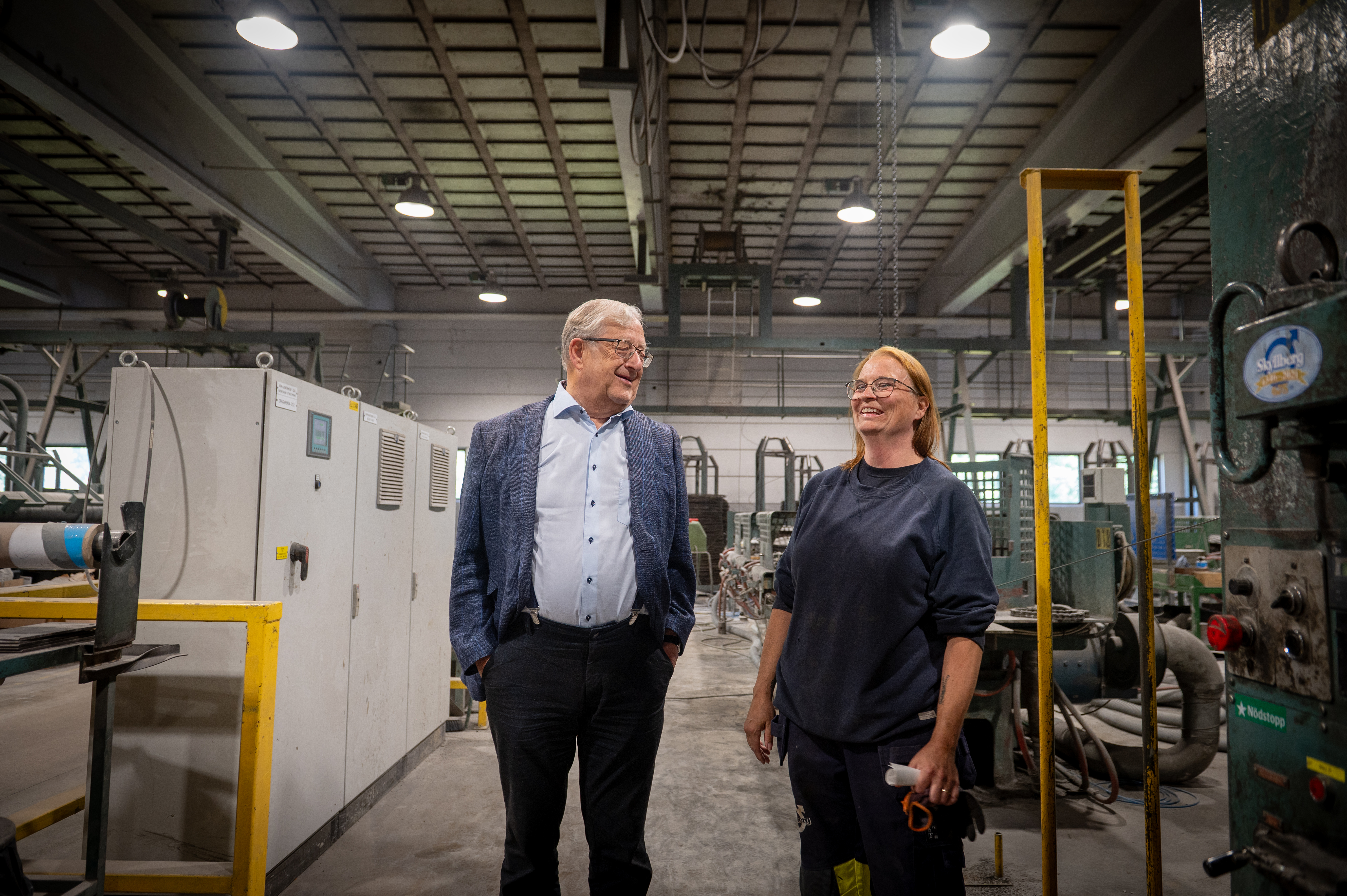 Gustaf Svensson och Linda Andersson står och pratar i en fabrik, omgivna av maskiner och utrustning under takbelysning.