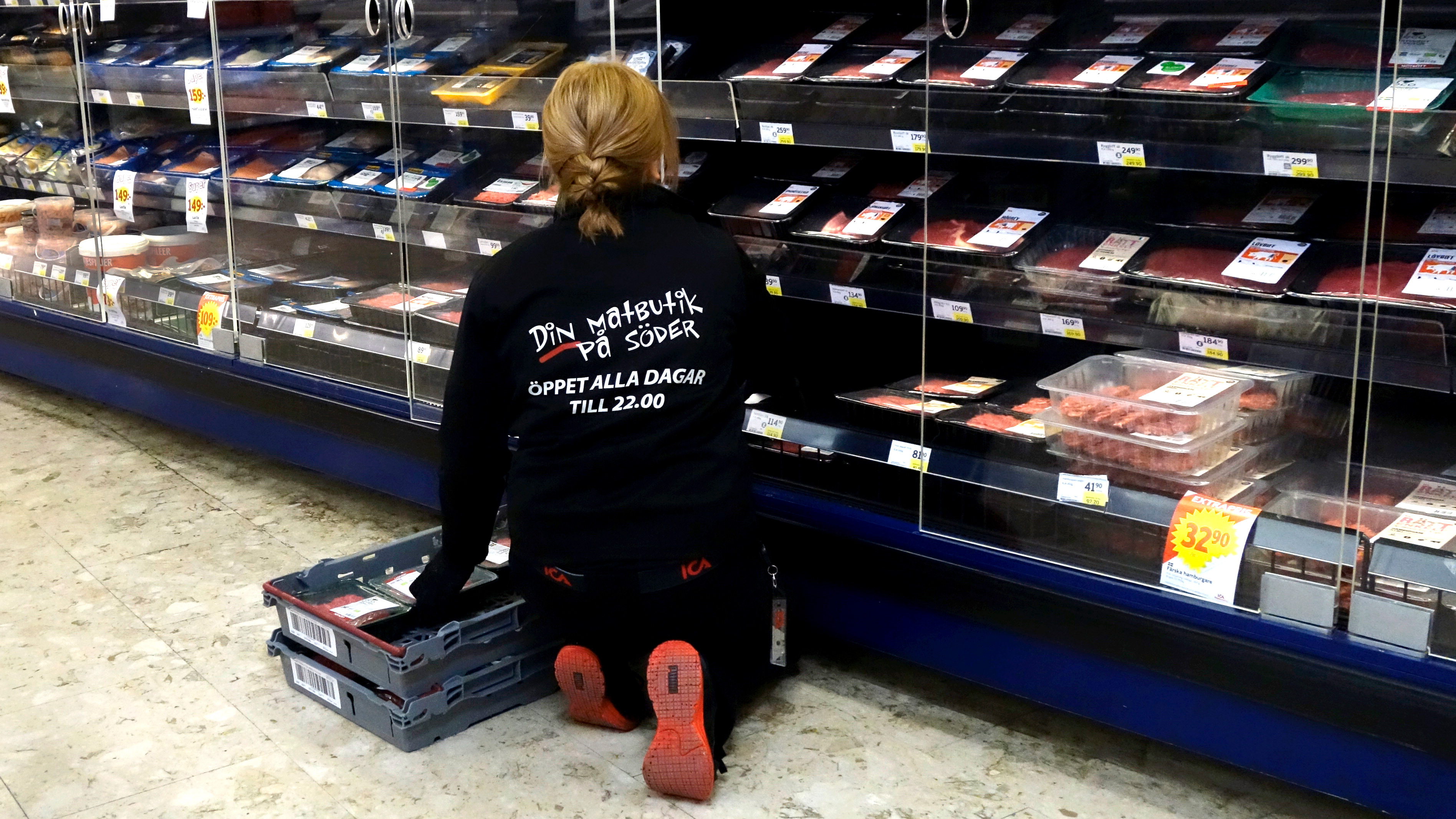 En butiksmedarbetare sitter på knä och fyller på förpackat kött i en kyldisk. Två lådor står intill.