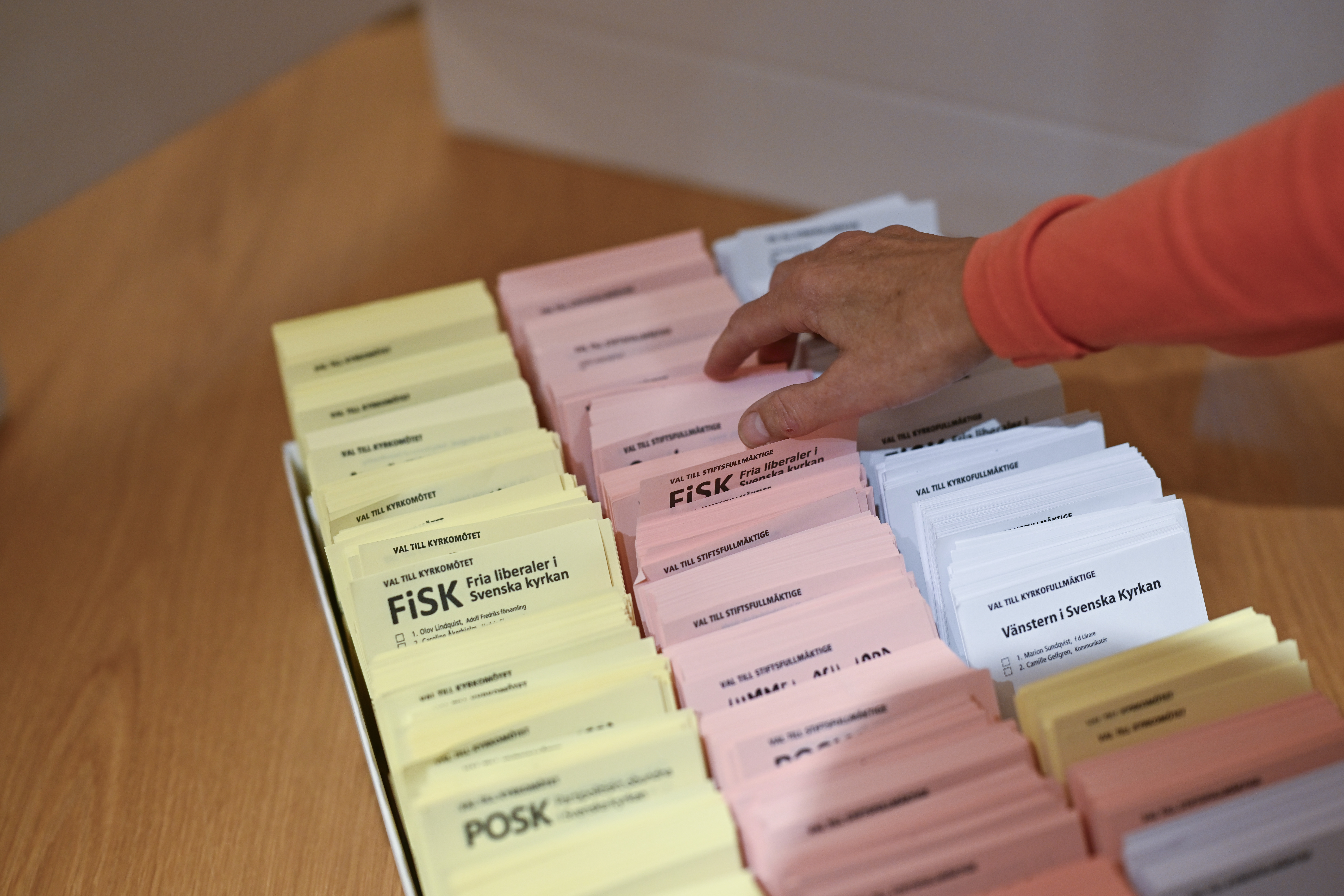 En person tar ett röstkort från staplar av gula, rosa och vita valsedlar som ligger på ett bord av trä.