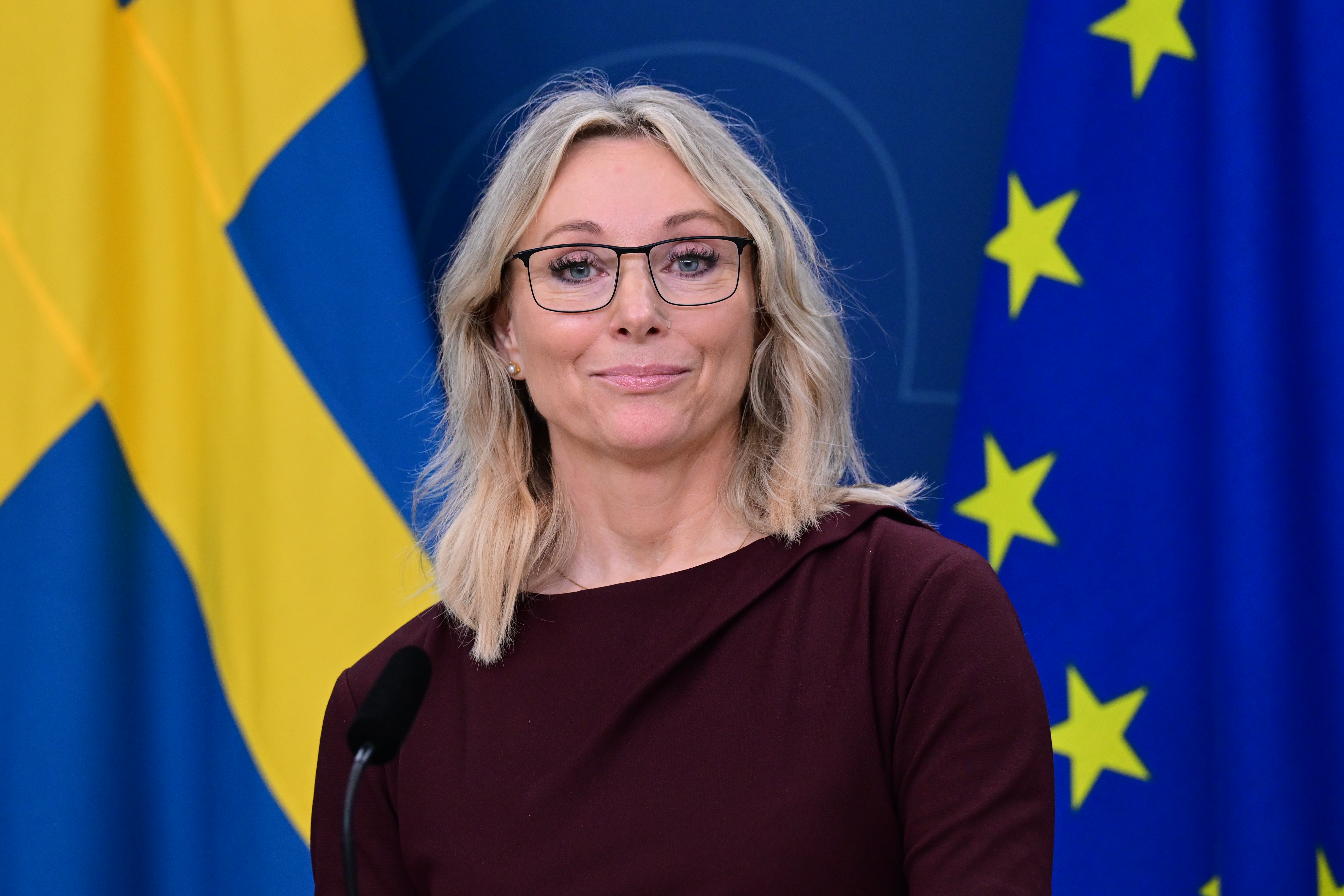 En kvinna med blont hår och glasögon står framför svenska och EU-flaggor och talar vid ett podium.