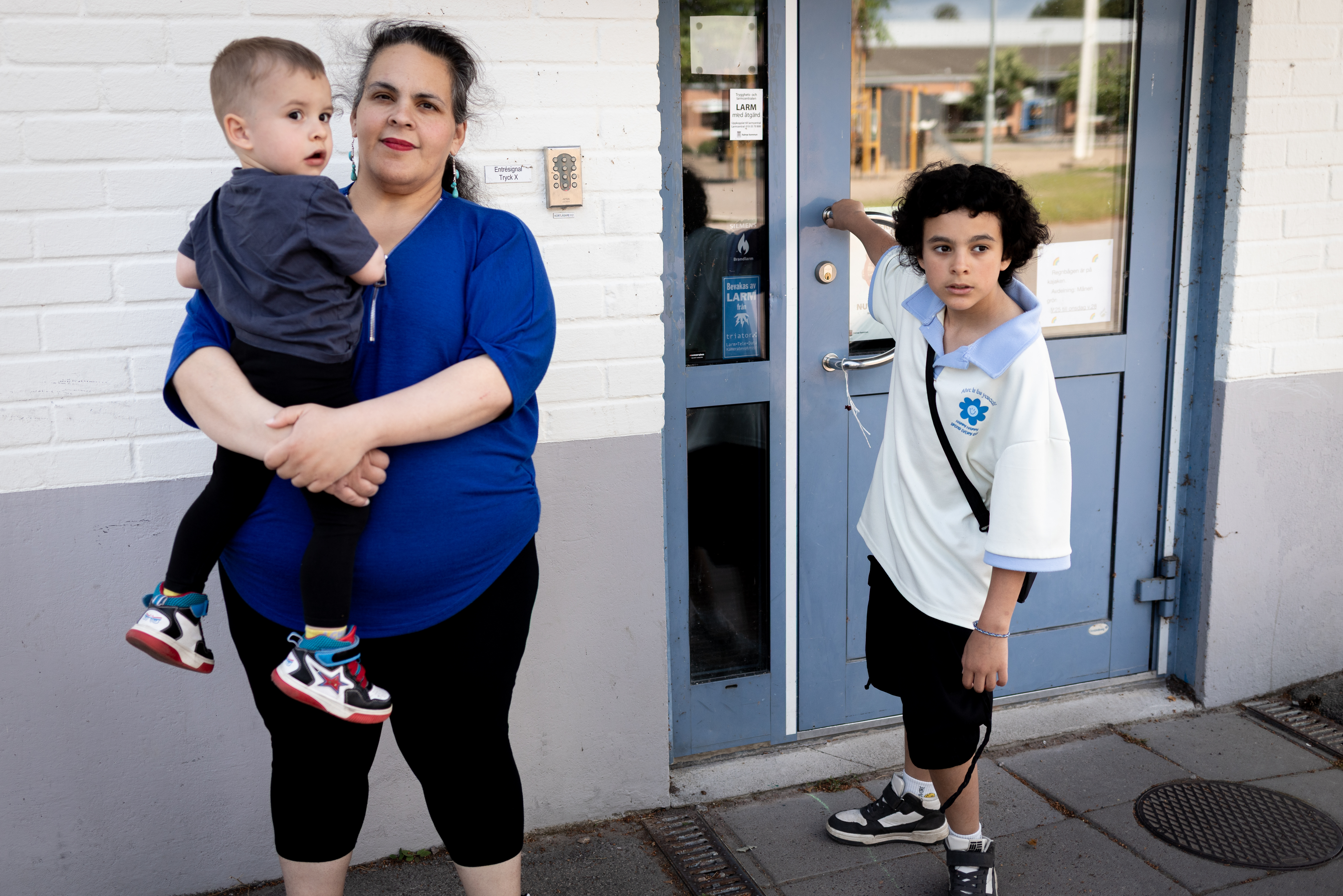 En kvinna med ett litet barn i famnen står bredvid en pojke som öppnar dörren till en byggnad.