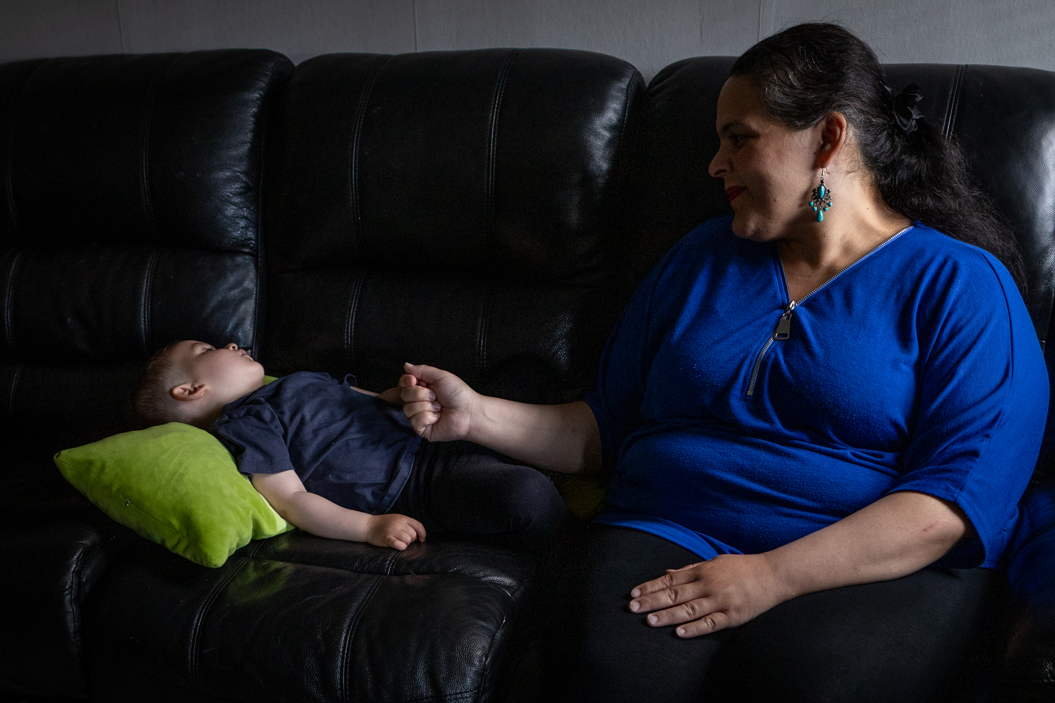 Anna Rashed i blå skjorta sitter i en soffa och håller handen på Robert som ligger med ett grönt kudd under huvudet.