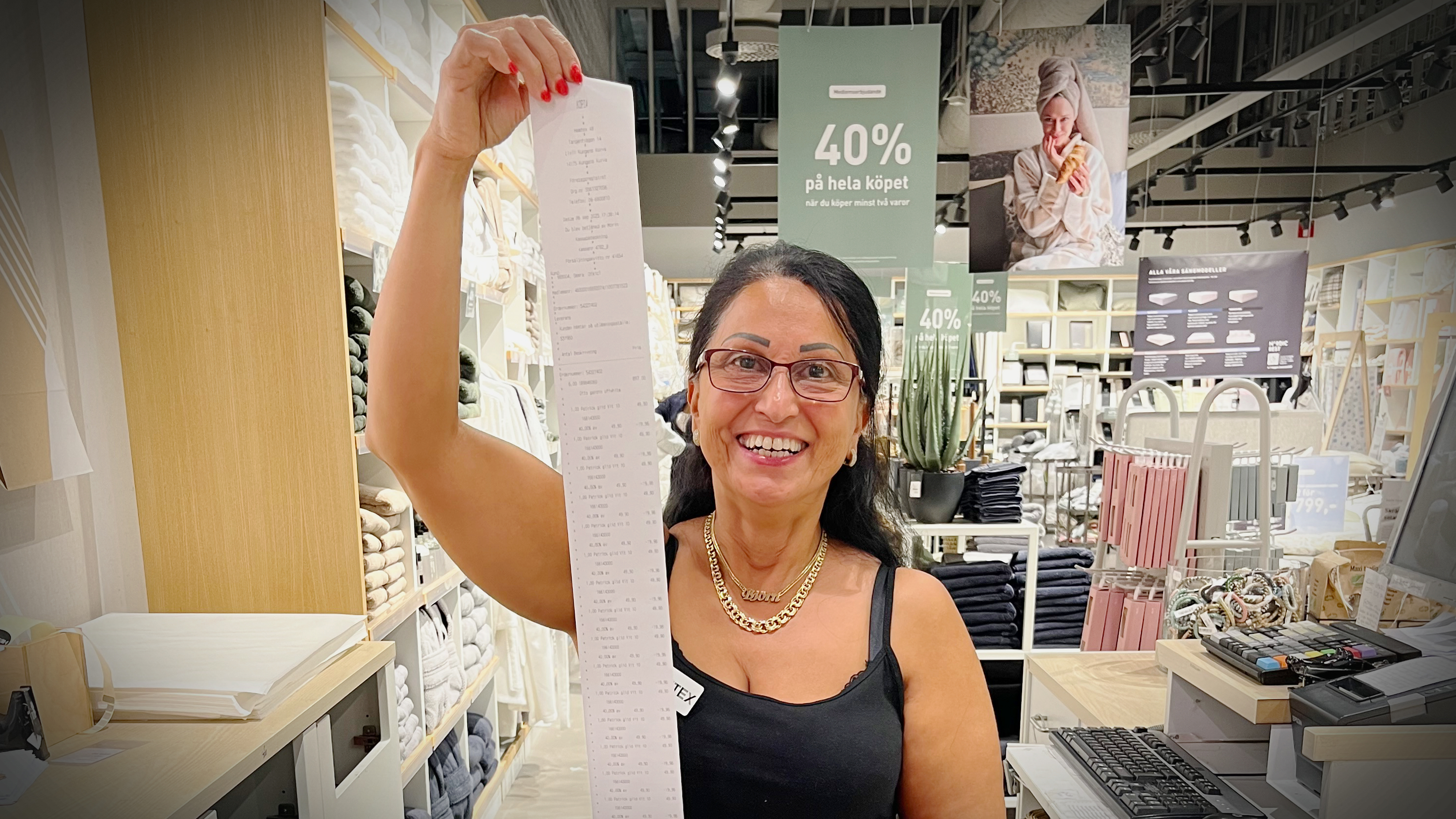 En kvinna står i en butik, håller upp ett långt kvitto bredvid en kassa. I bakgrunden finns reaskyltar och varor.