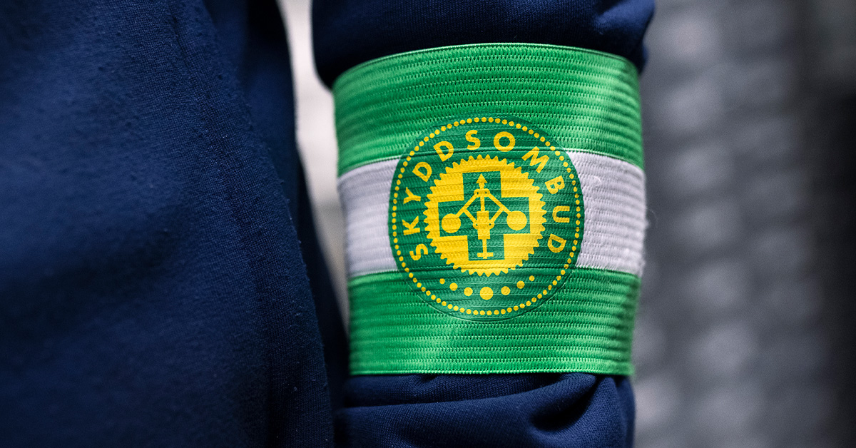 Ett grönt och vitt armband med en gul och blå rund symbol med texten "SKYDDSOMBUD" bärs på en mörkblå ärm.