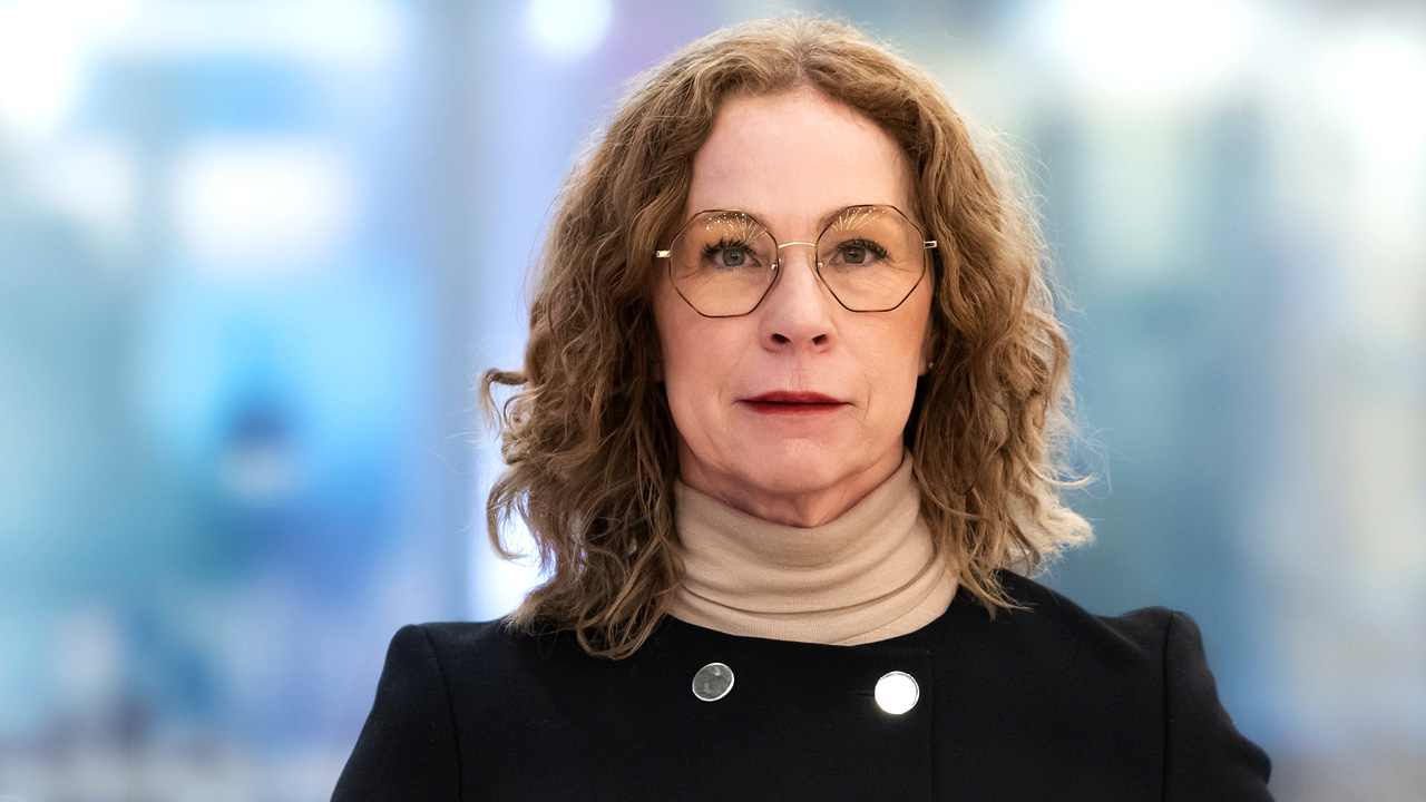 En person med lockigt hår, glasögon och en beige polotröja under en svart jacka står vänd mot kameran framför suddig bakgrund.