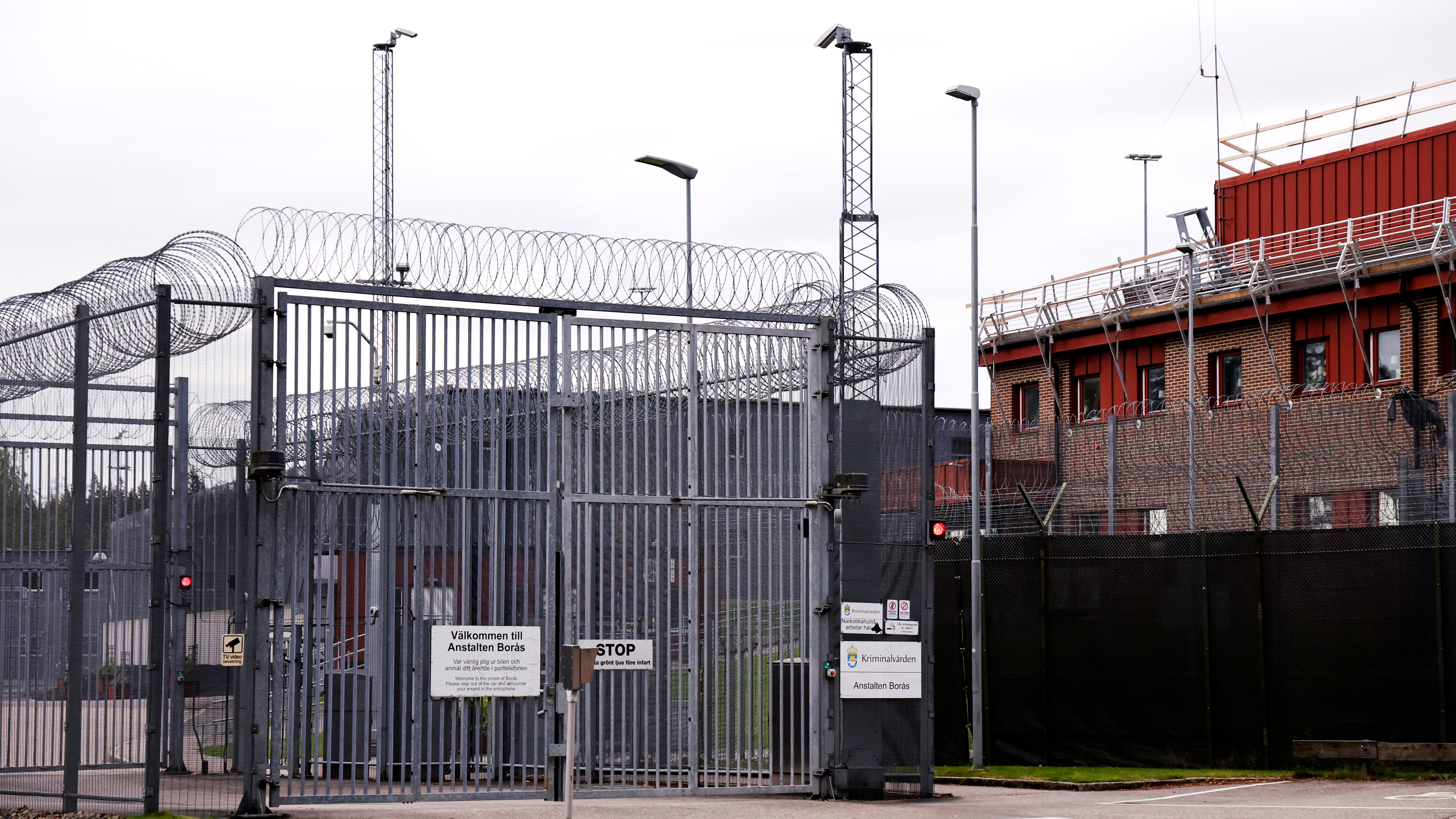 En anläggning med höga stängsel, taggtråd, övervakningskameror och varningsskyltar vid grind intill en tegelbyggnad.