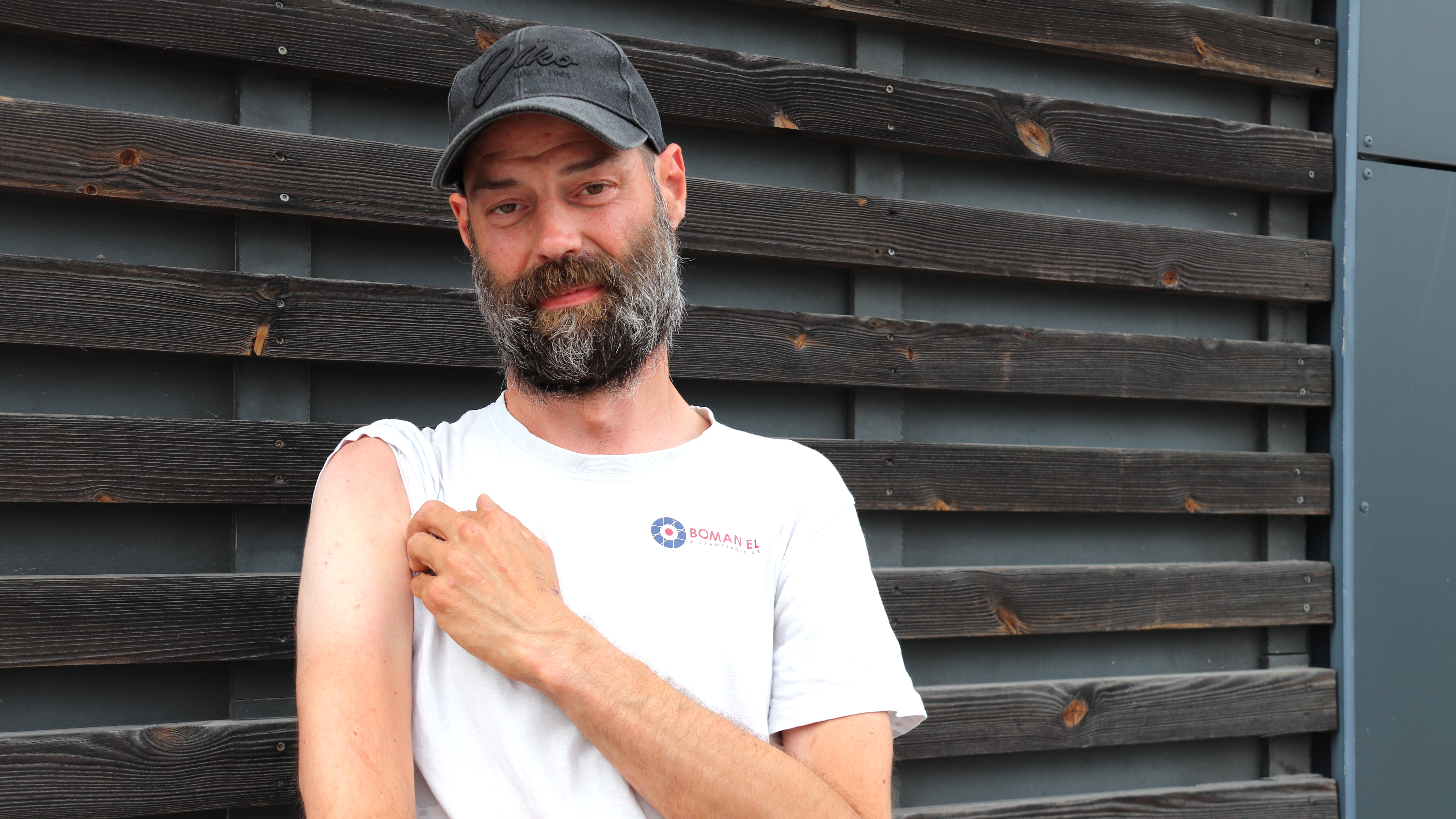 En man med vit t-shirt och svart keps står framför en trävägg och lyfter ärmen för att visa sin överarm.