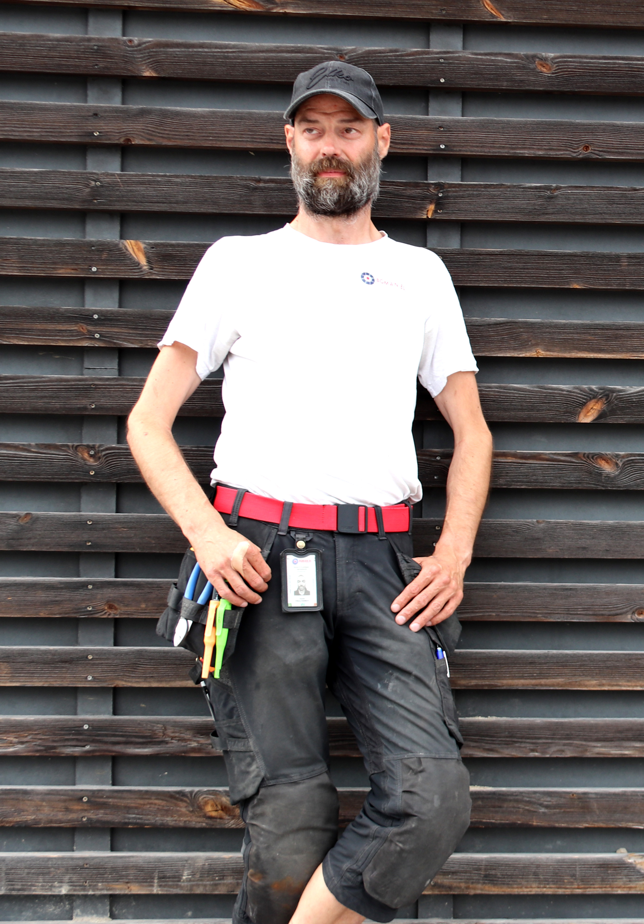 En man med vit skjorta, svarta byxor med verktyg och svarta kepsar står mot en trävägg och tittar lite åt sidan.