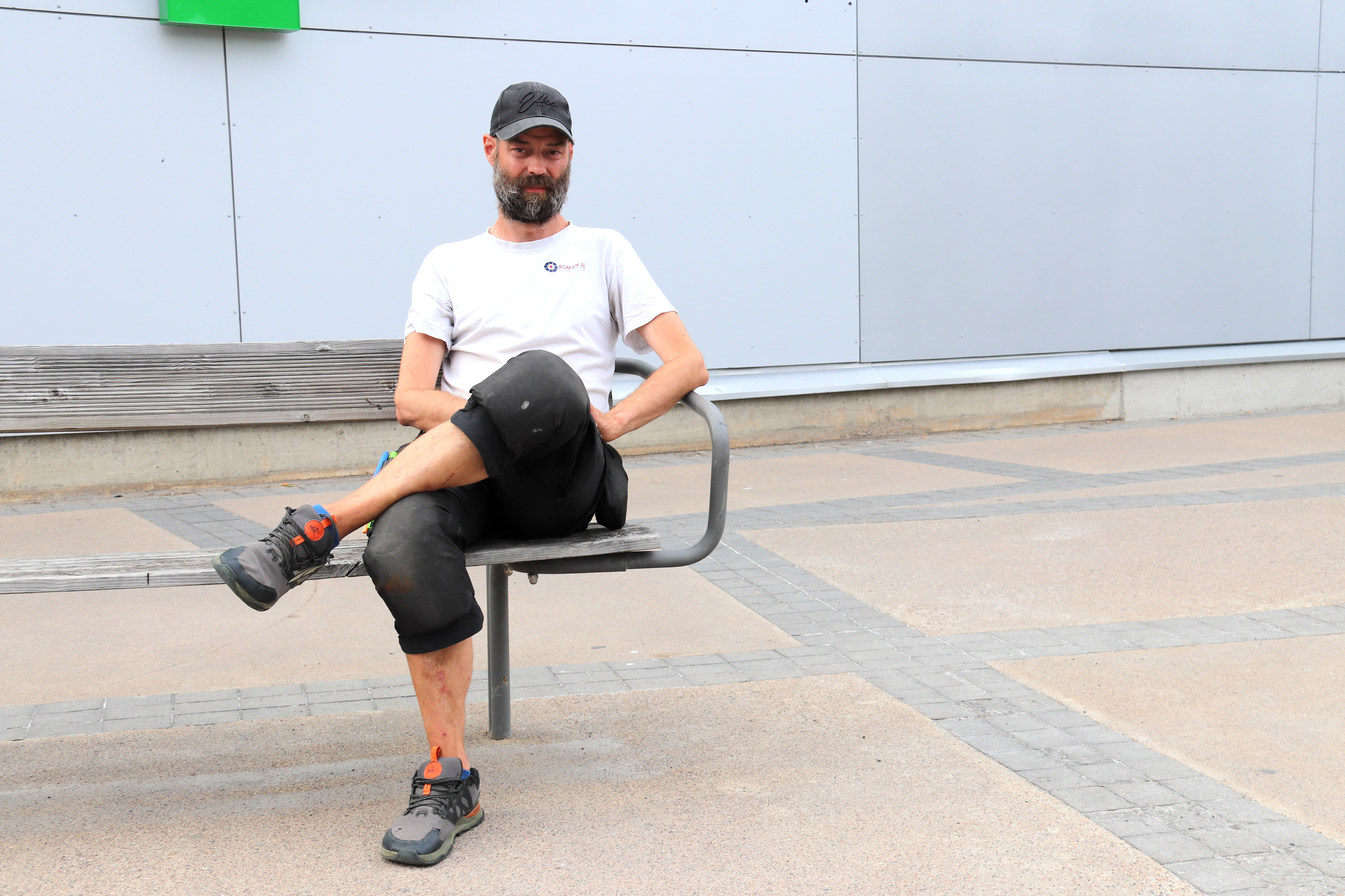 En man med skägg, vit t-shirt, svarta kepsar och svarta byxor sitter med korslagda ben på en metallbänk utomhus.