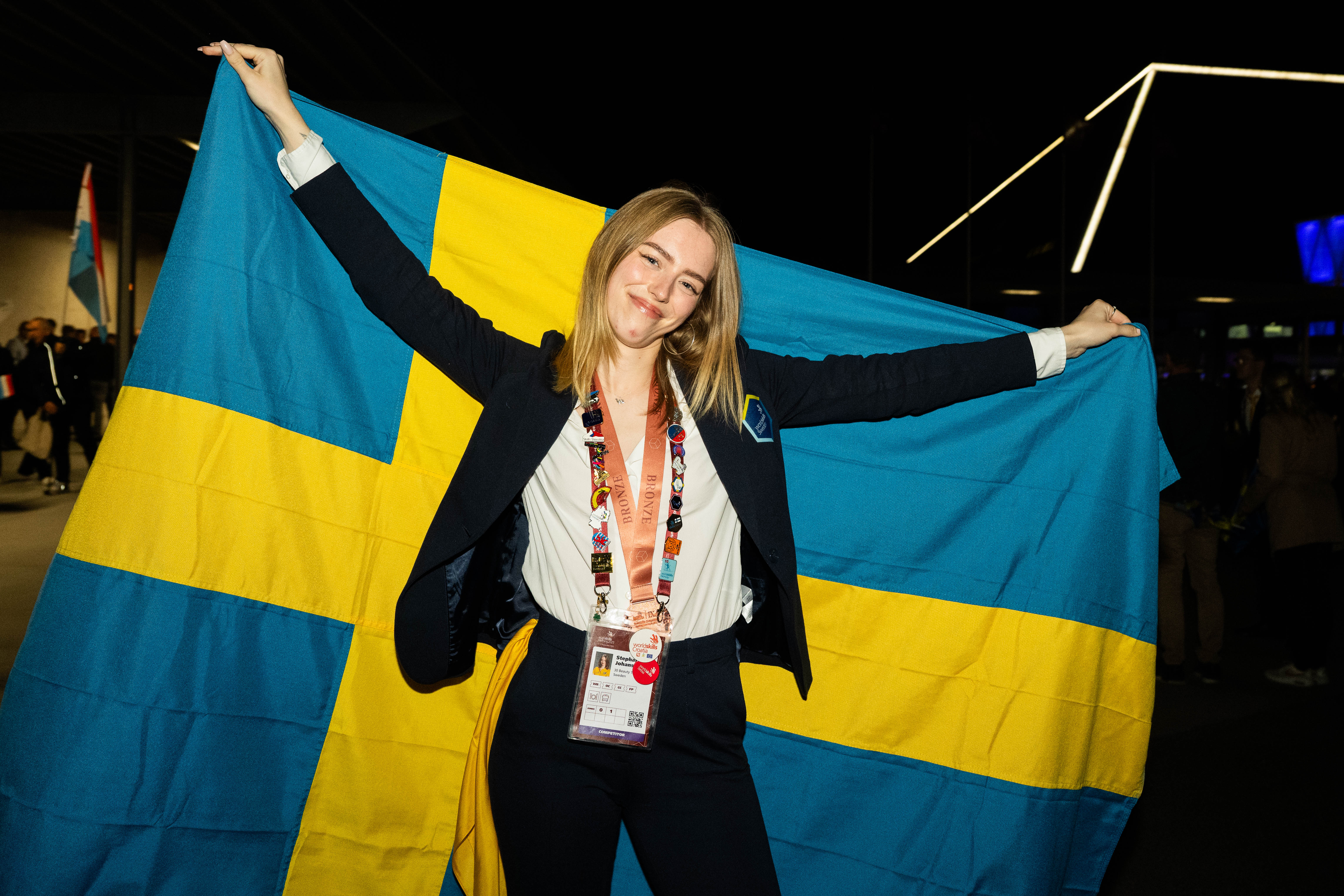 En person står med en stor svensk flagga bakom ryggen, klädd i kostym och evenemangsband, med folk och lampor i bakgrunden.