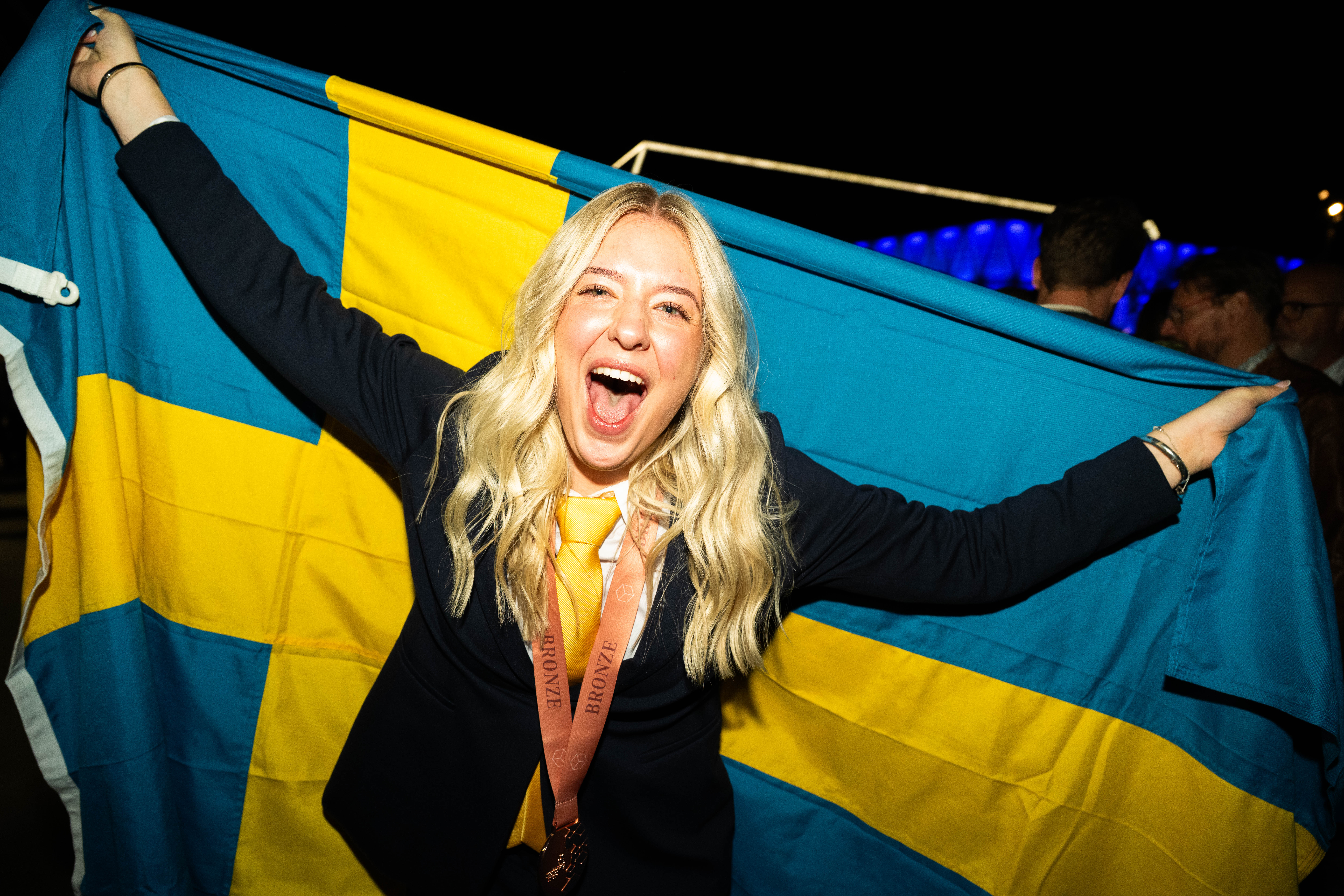 En person med långt blont hår håller upp en svensk flagga, ler med öppen mun och har kostym och en bronsmedalj runt halsen.