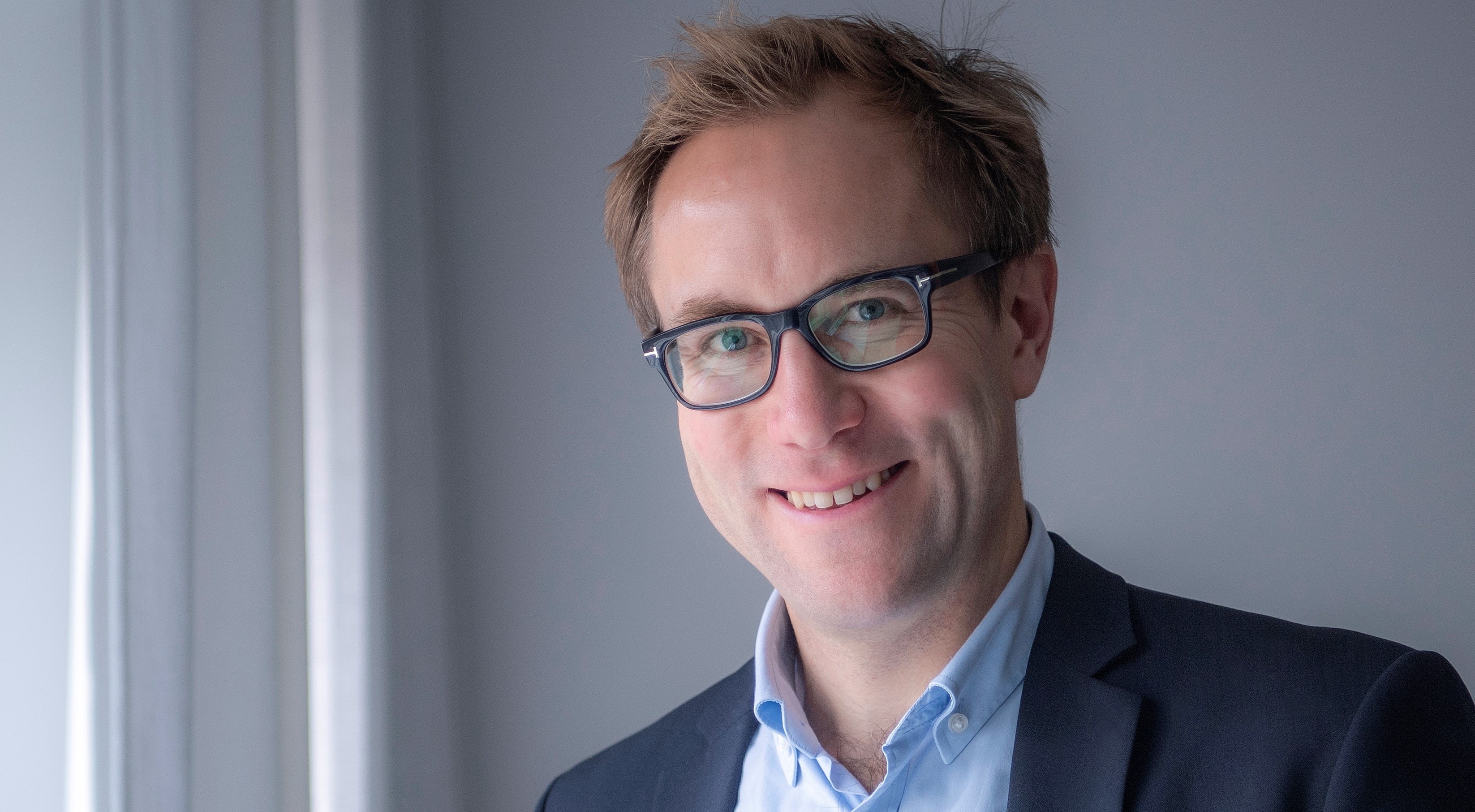 En man med glasögon och en mörk blå kavaj över en ljusblå skjorta står inomhus nära ett fönster och ler.