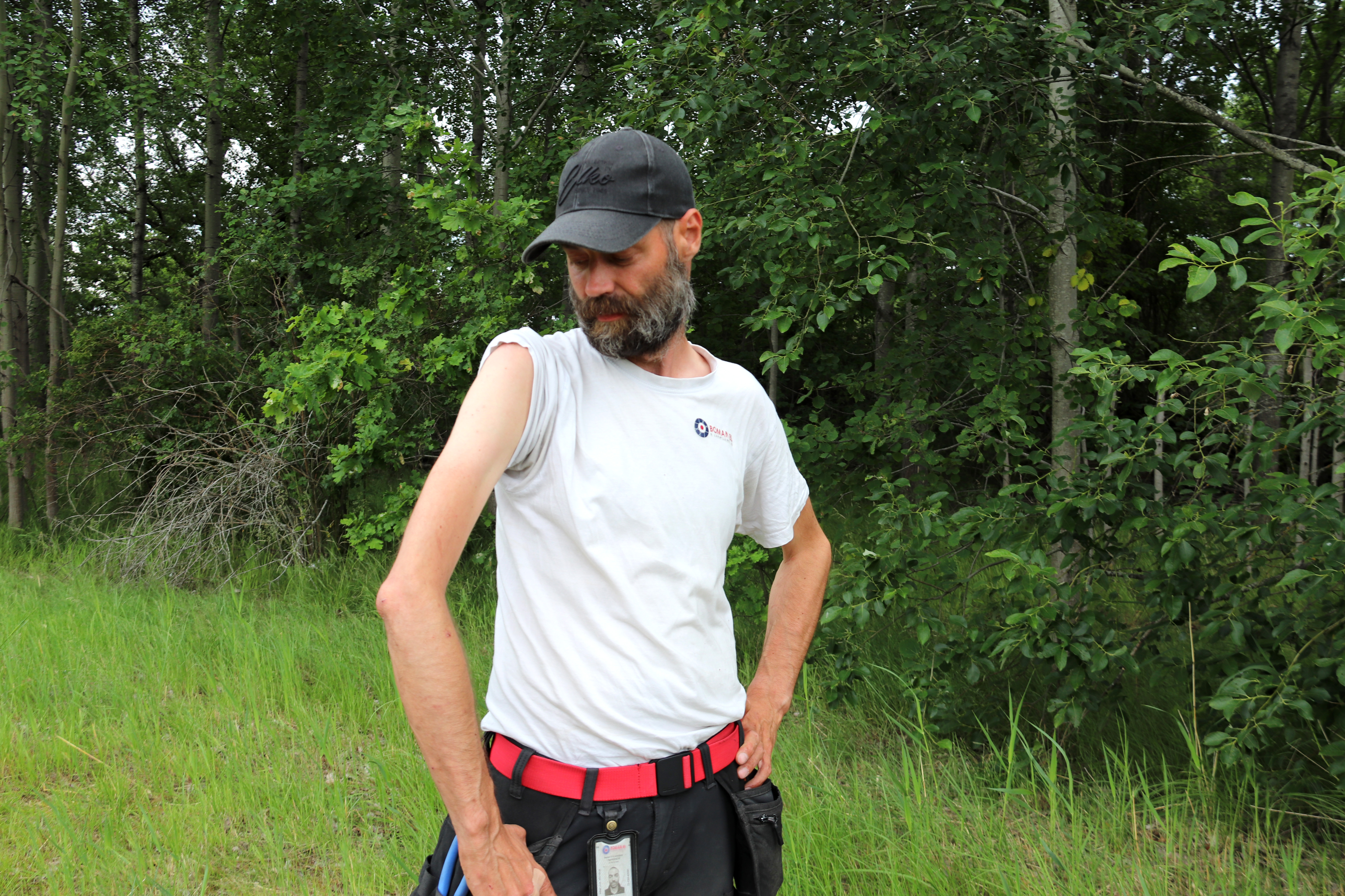 En man med vit ärmlös tröja och svart keps står utomhus framför träd och gräs och tittar ner på sin vänstra arm.