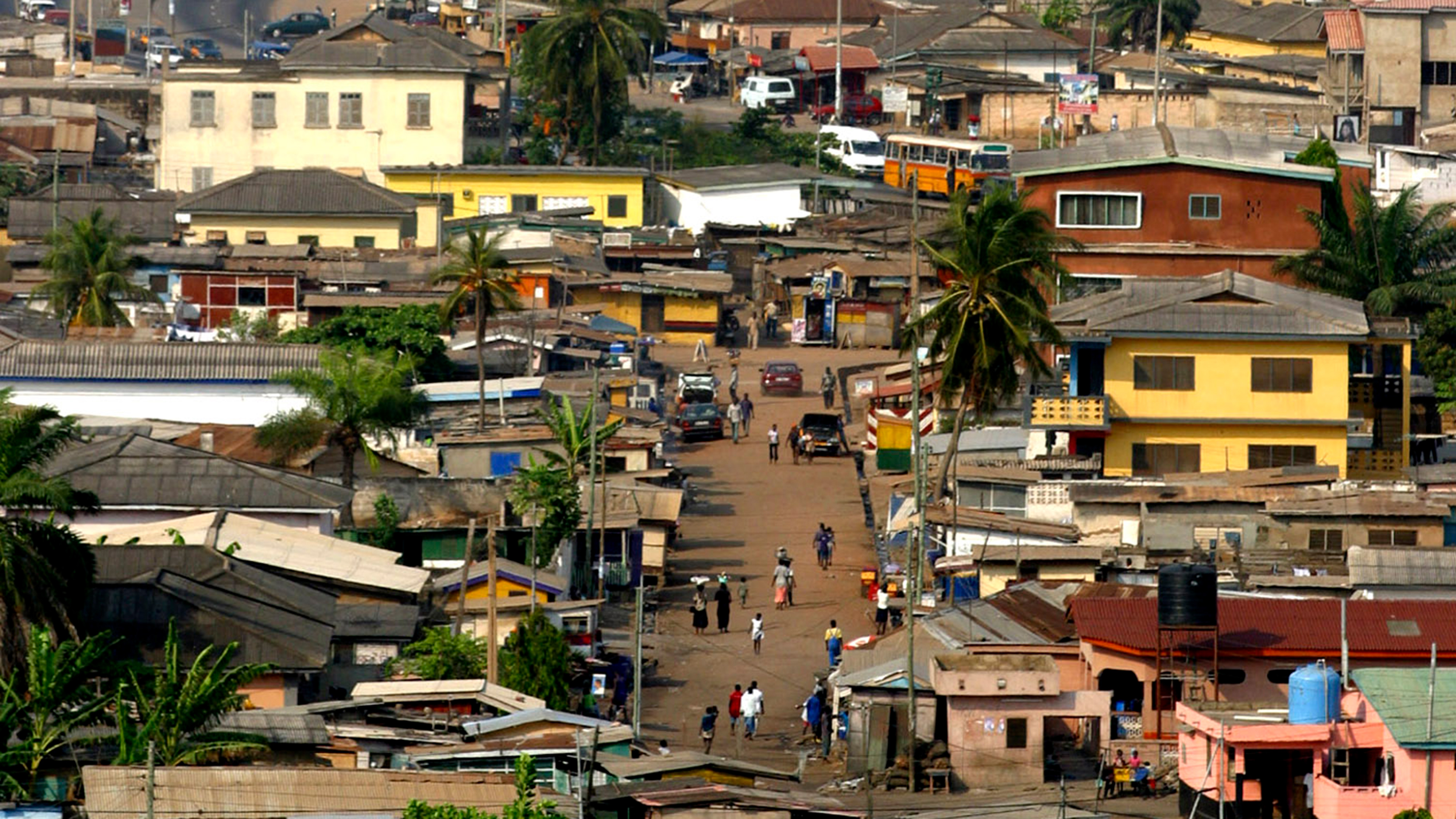 En gata i ett stadsområde med människor som går, parkerade bilar och både bostadshus och affärsbyggnader under palmer.