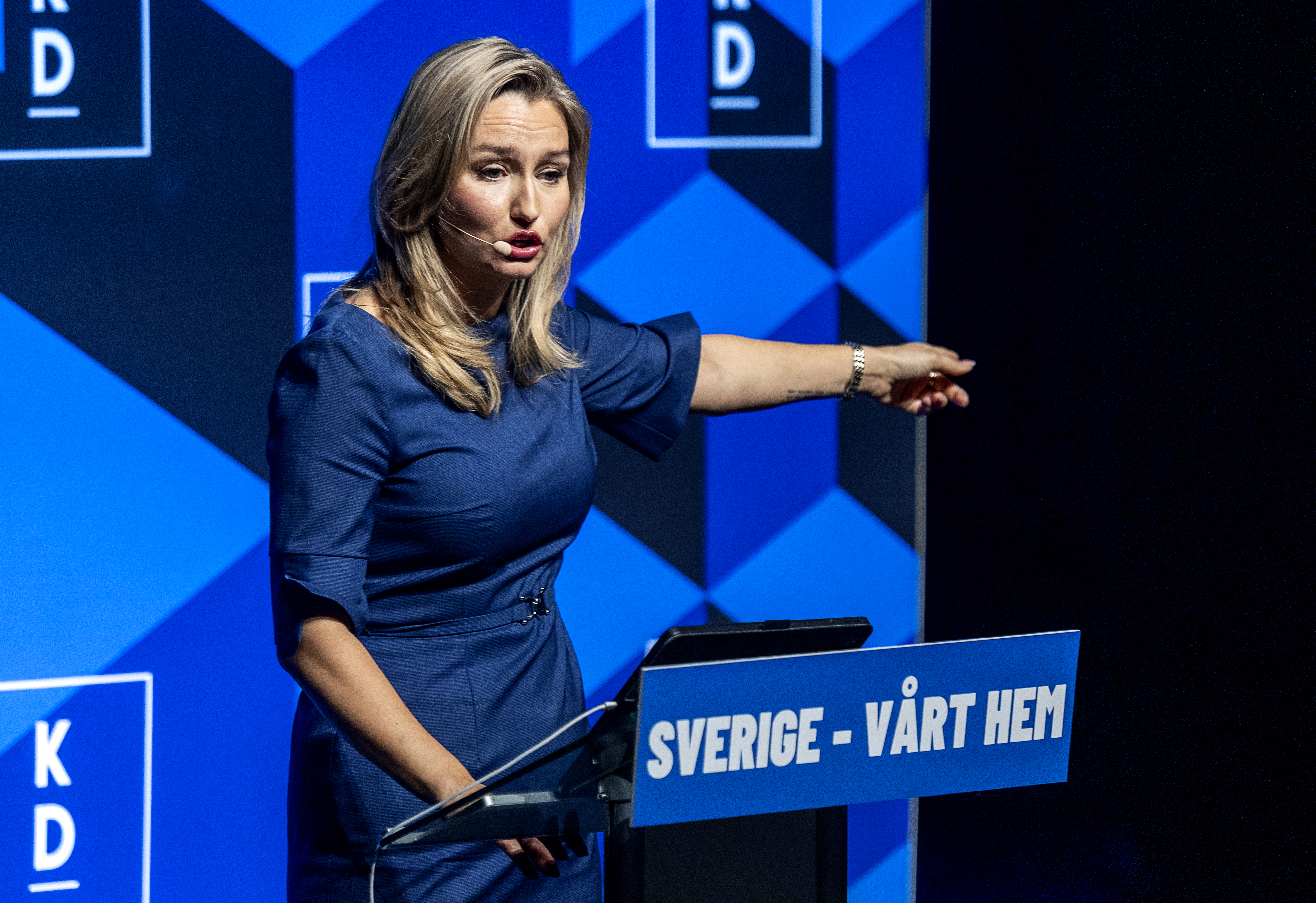 En kvinna i blå klänning talar vid ett podium med skylten "Sverige - Vårt Hem" och pekar till höger mot blå bakgrund.
