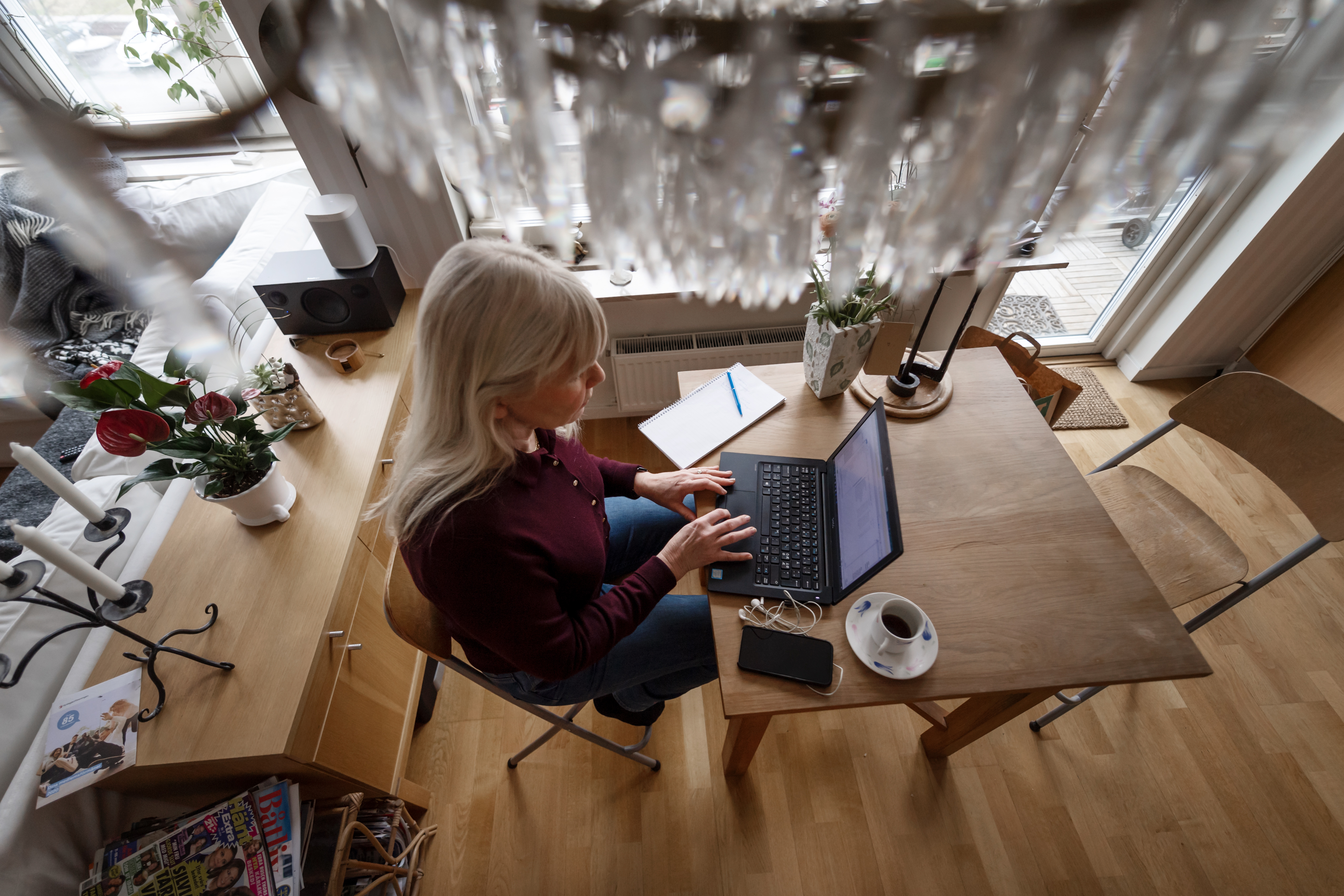En kvinna sitter vid ett bord och använder en laptop med en anteckningsbok, en mobiltelefon och en kaffekopp bredvid sig.