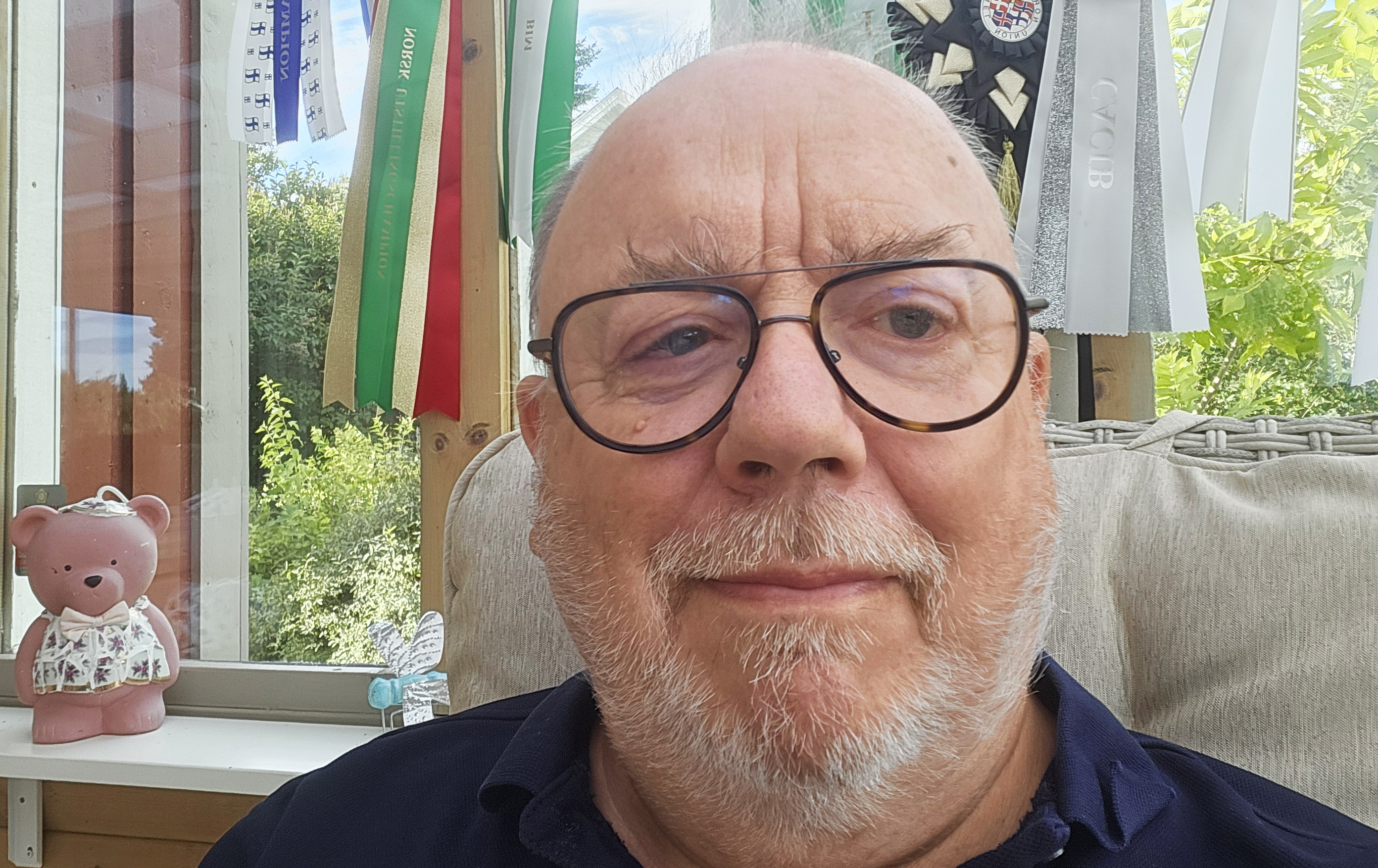 En man med glasögon och skägg sitter inomhus nära ett fönster.