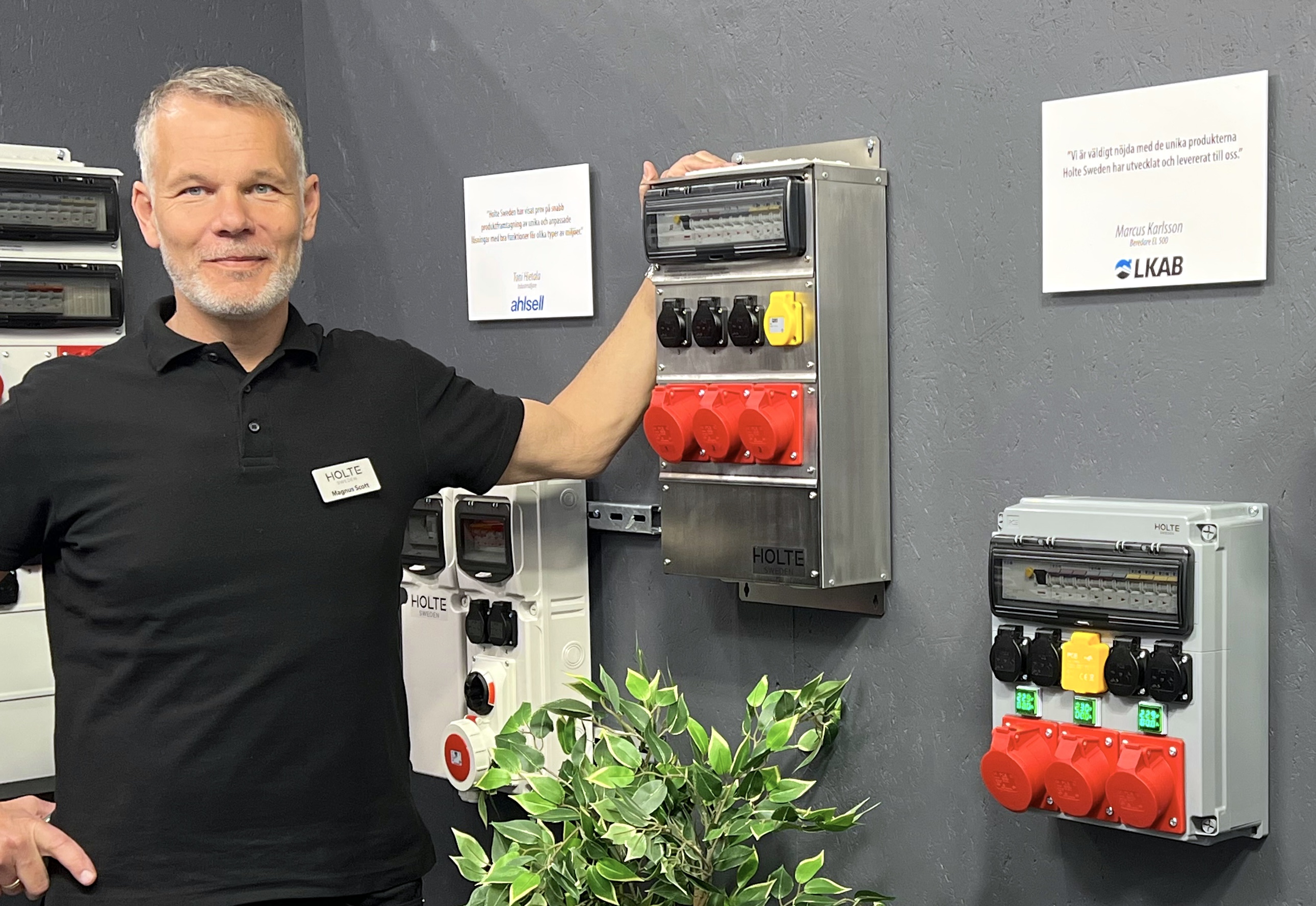 En man står bredvid industriella elpaneler som är monterade på en vägg. Informationsskyltar och en krukväxt syns också.