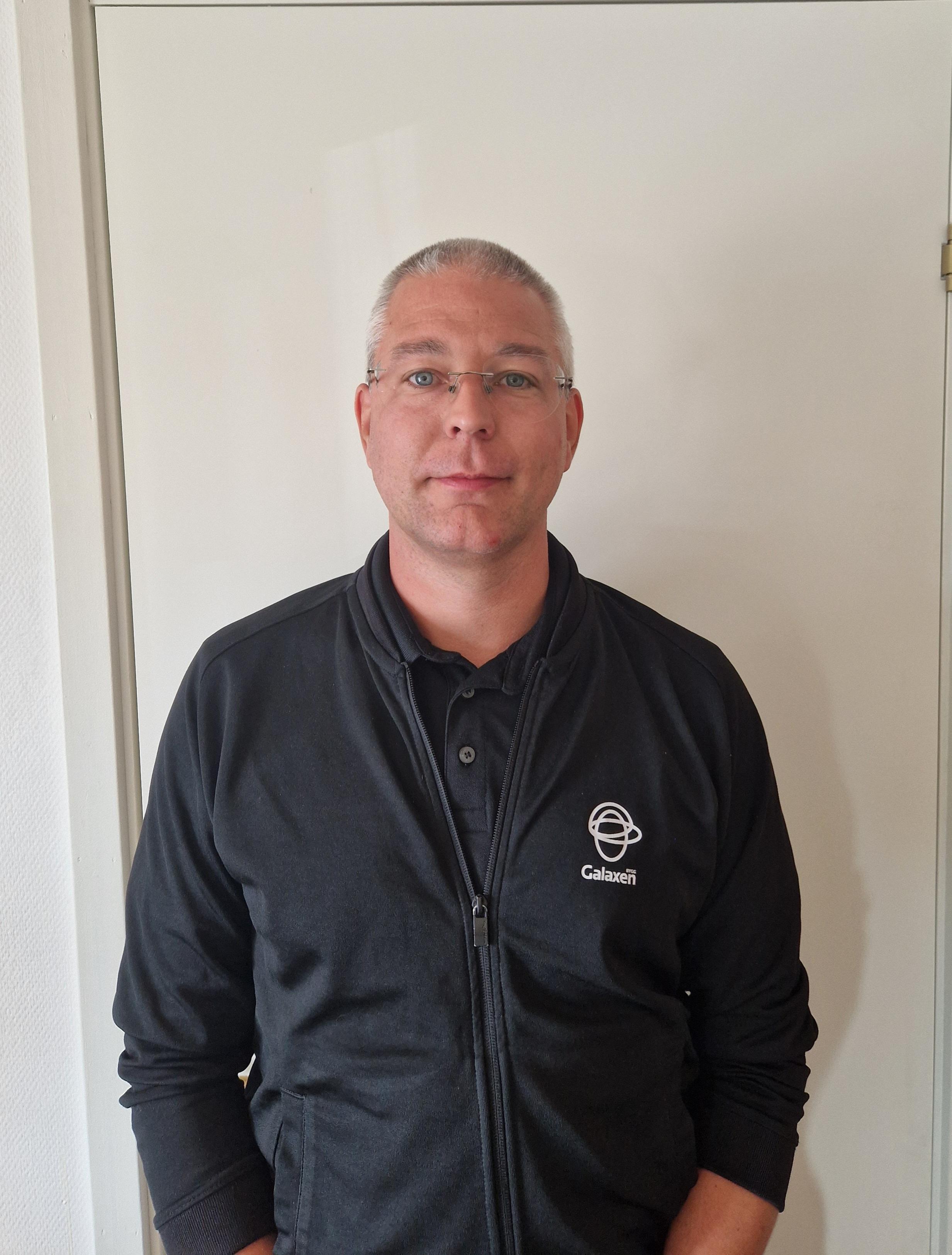 En man med kort grått hår och glasögon står framför en vit dörr, iförd en svart jacka med "Galaxenti"-logga och svart skjorta.