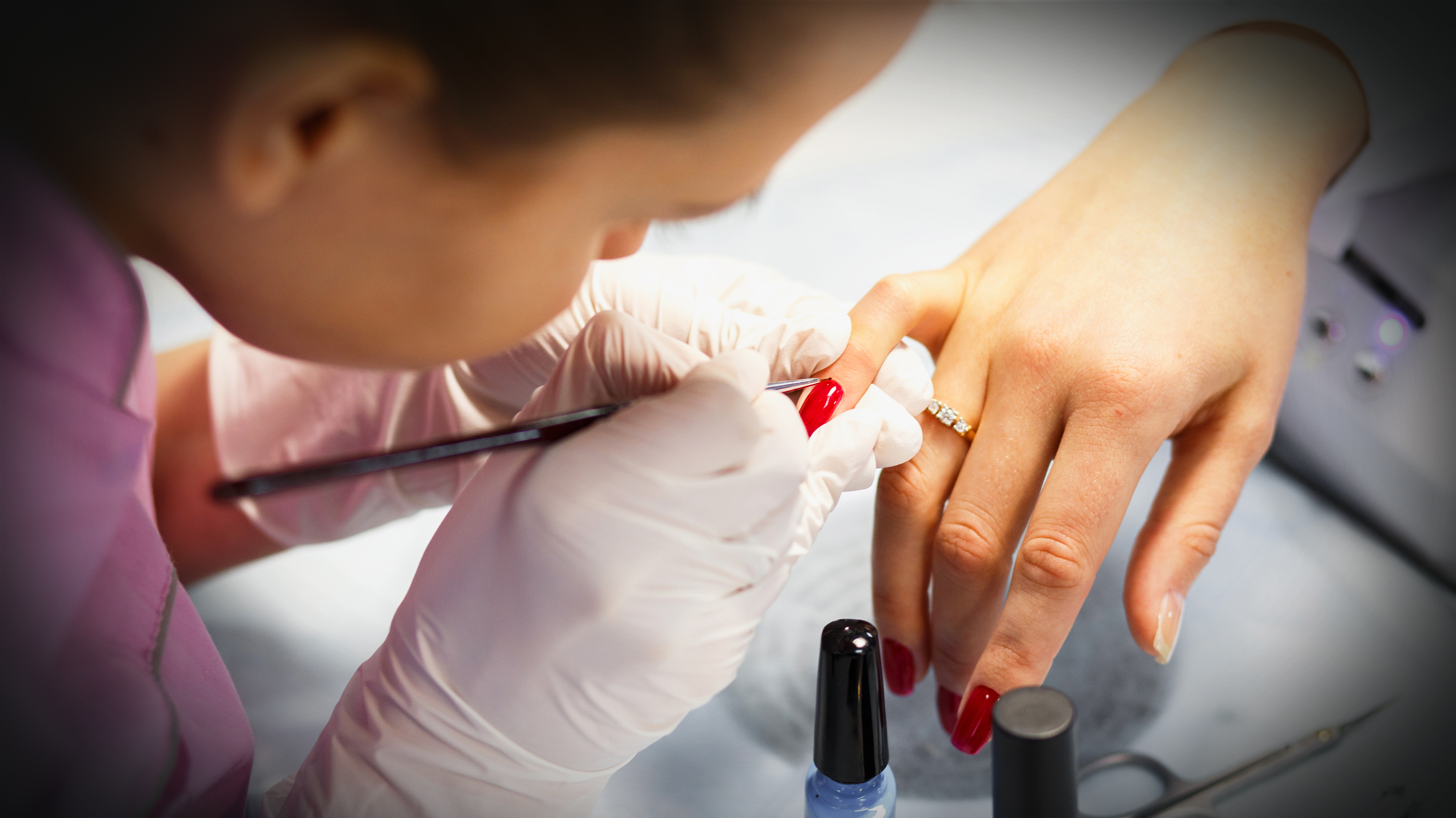 En person med handskar målar röd nagellack på en annans naglar, som bär en ring. Nagellacksflaskor står på bordet.