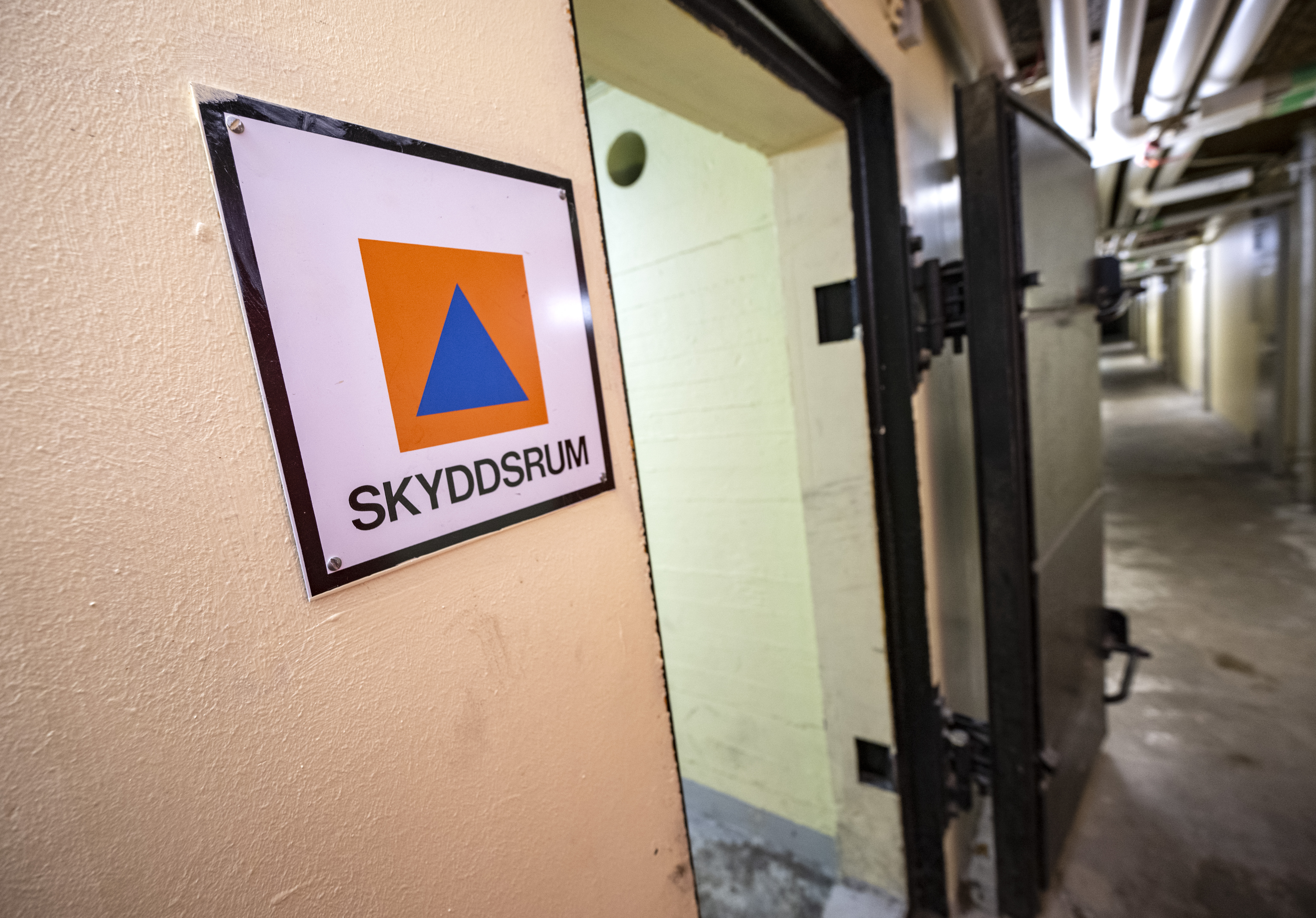 En dörr i en korridor med en skylt där det står "Skyddsrum" och en orangeblå triangel som markerar ett svenskt skyddsrum.