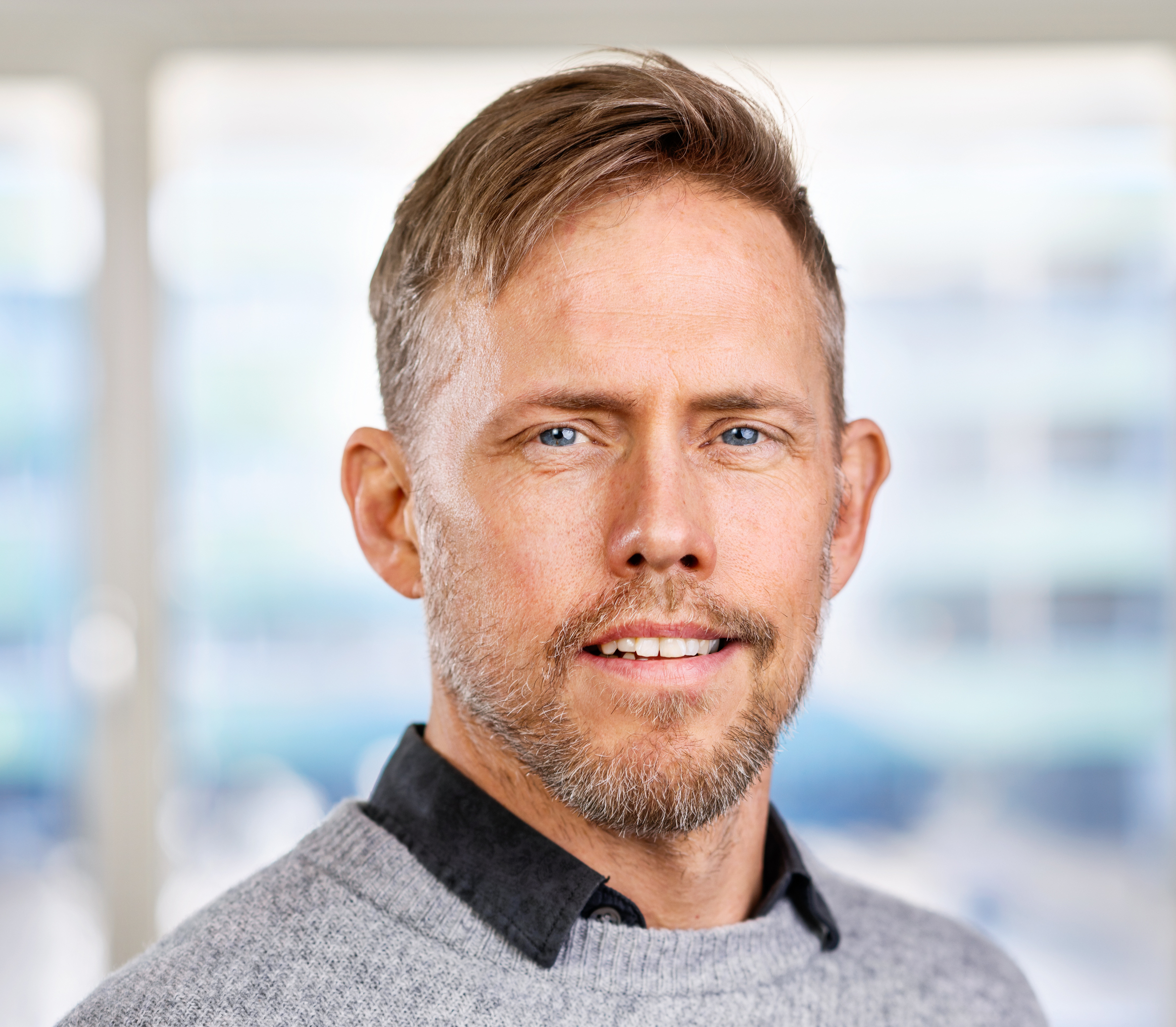 En man med kort ljusbrunt hår, skägg och blå ögon har en grå tröja över en svart skjorta och står inomhus.